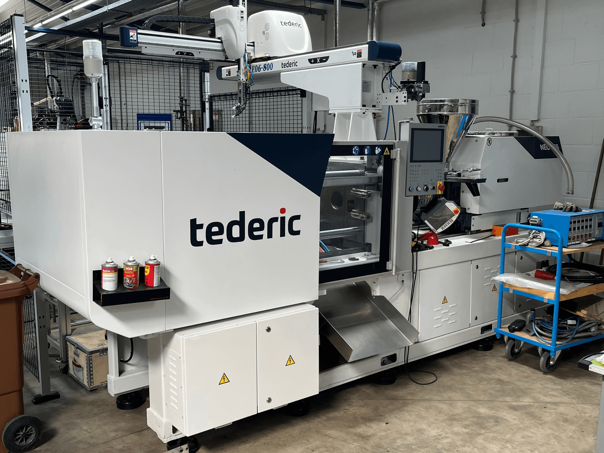 Bir atölyede bulunan TEDERIC NEO.E55/e110h hidrolik enjeksiyon kalıplama makinesi; kontrol paneli ve yanındaki aletlerin görüldüğü yandan görünüm.