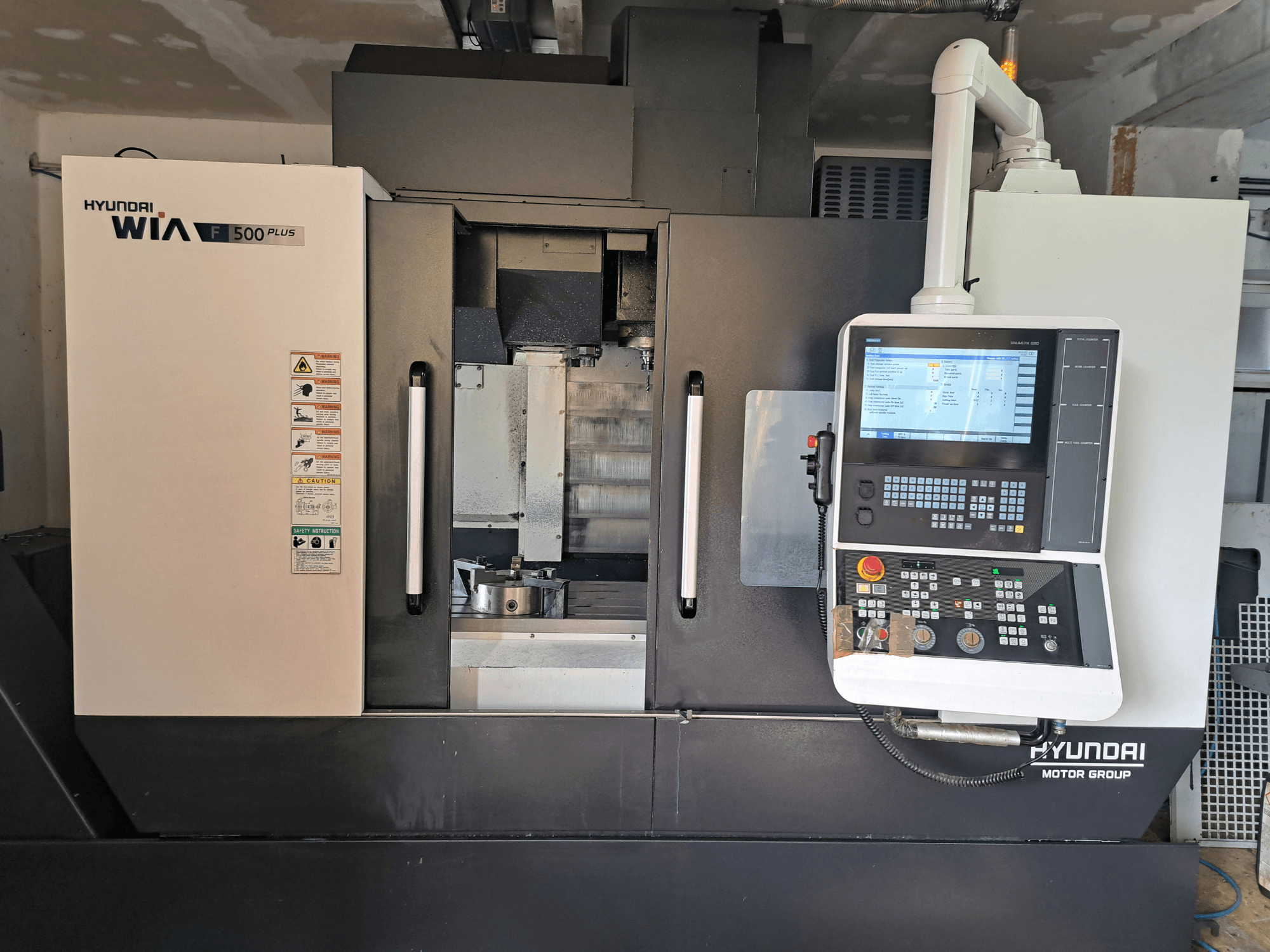 Hyundai WIA F500 Plus CNC makinesinin önden görünümü, dijital kontrol paneli ve güvenlik etiketleri.