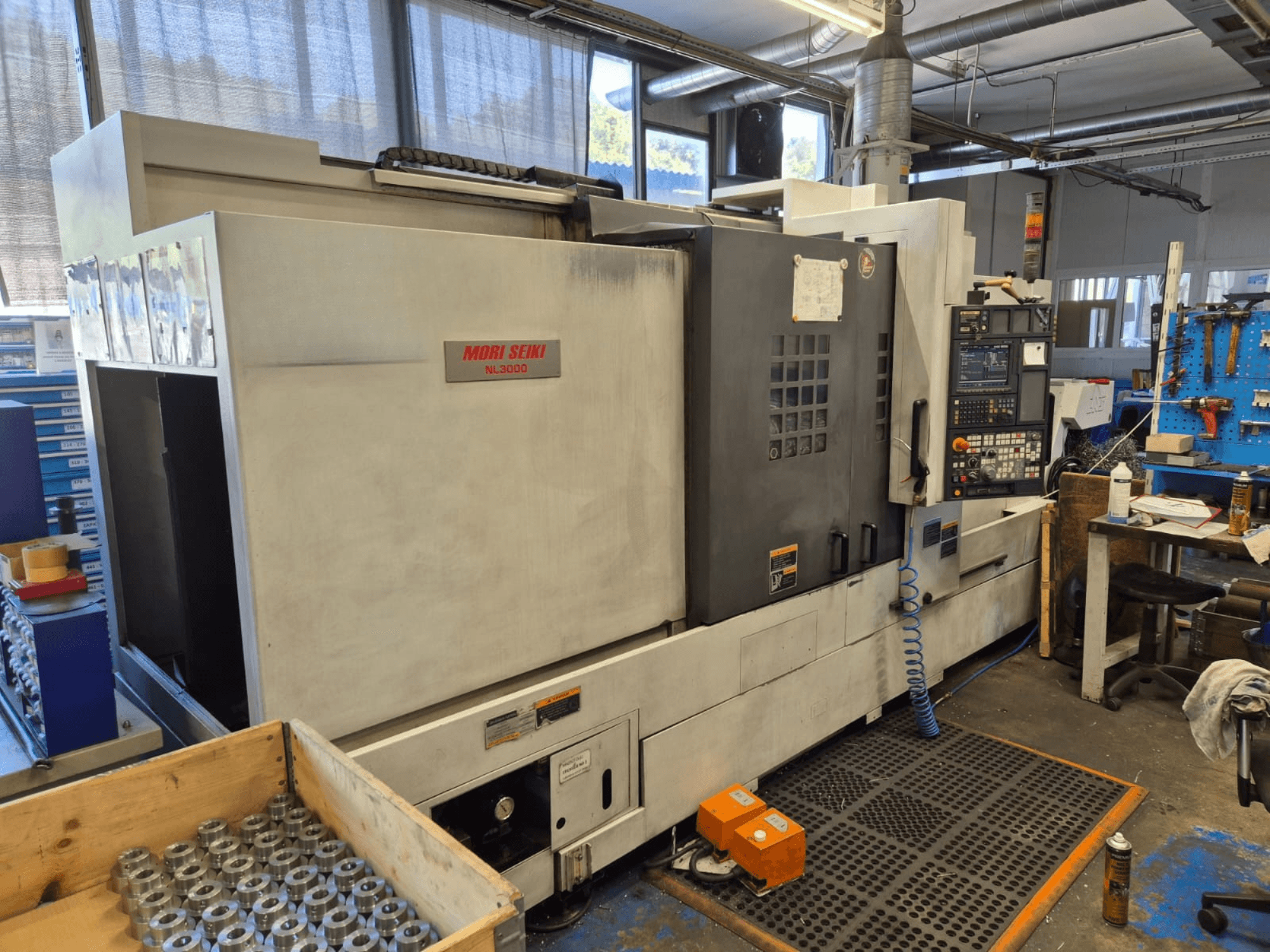 Bir atölyede Mori Seiki NL3000 CNC torna tezgahı, yandan görünümde kontrol paneli, aletler ve ahşap bir kutu içinde metal parçalar sergileniyor.