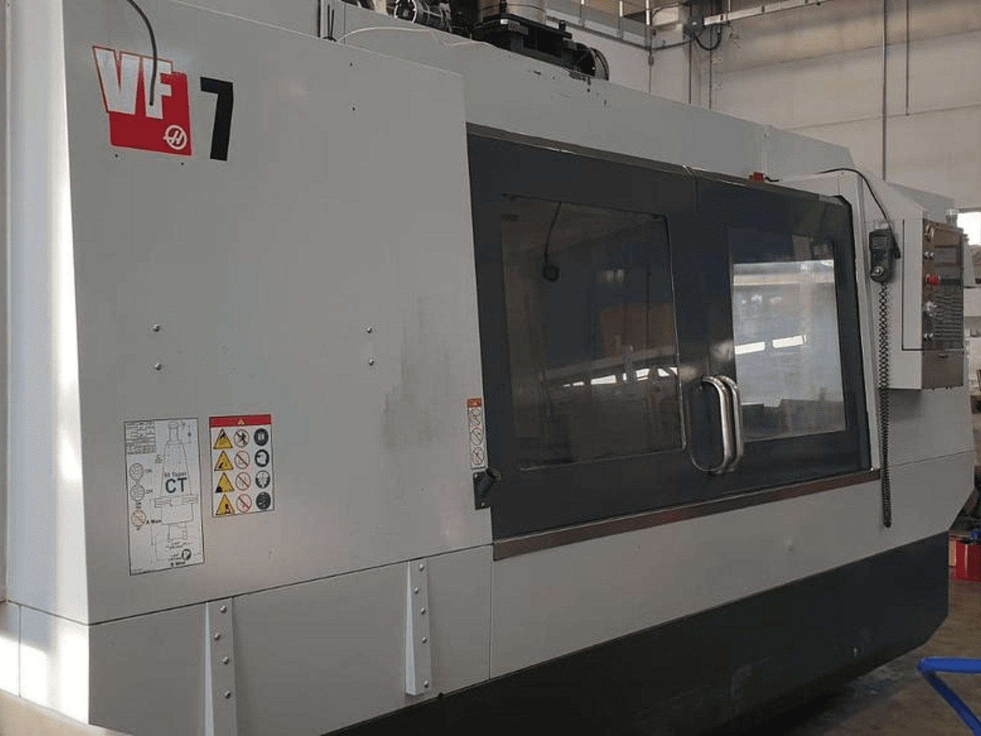 Haas VF-7 CNC makinesi yandan görünümde, metal gri kaplama, kontrol paneli ve güvenlik etiketlerini sergiliyor.