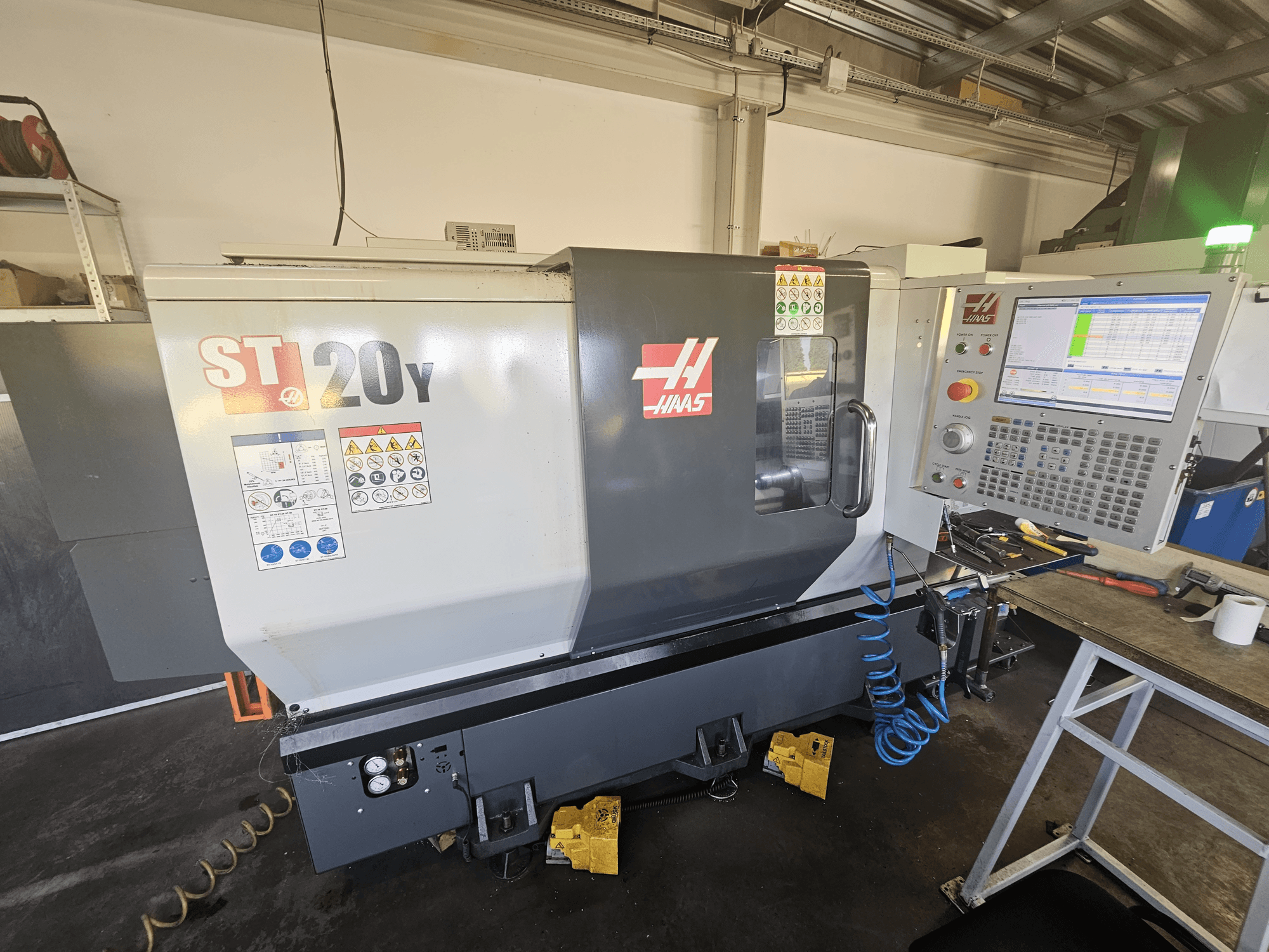 Haas ST-20Y CNC torna tezgahı, kontrol paneli ile önden görünüm; güvenlik etiketleri ve yan tarafta hava hortumu detayı içerir.
