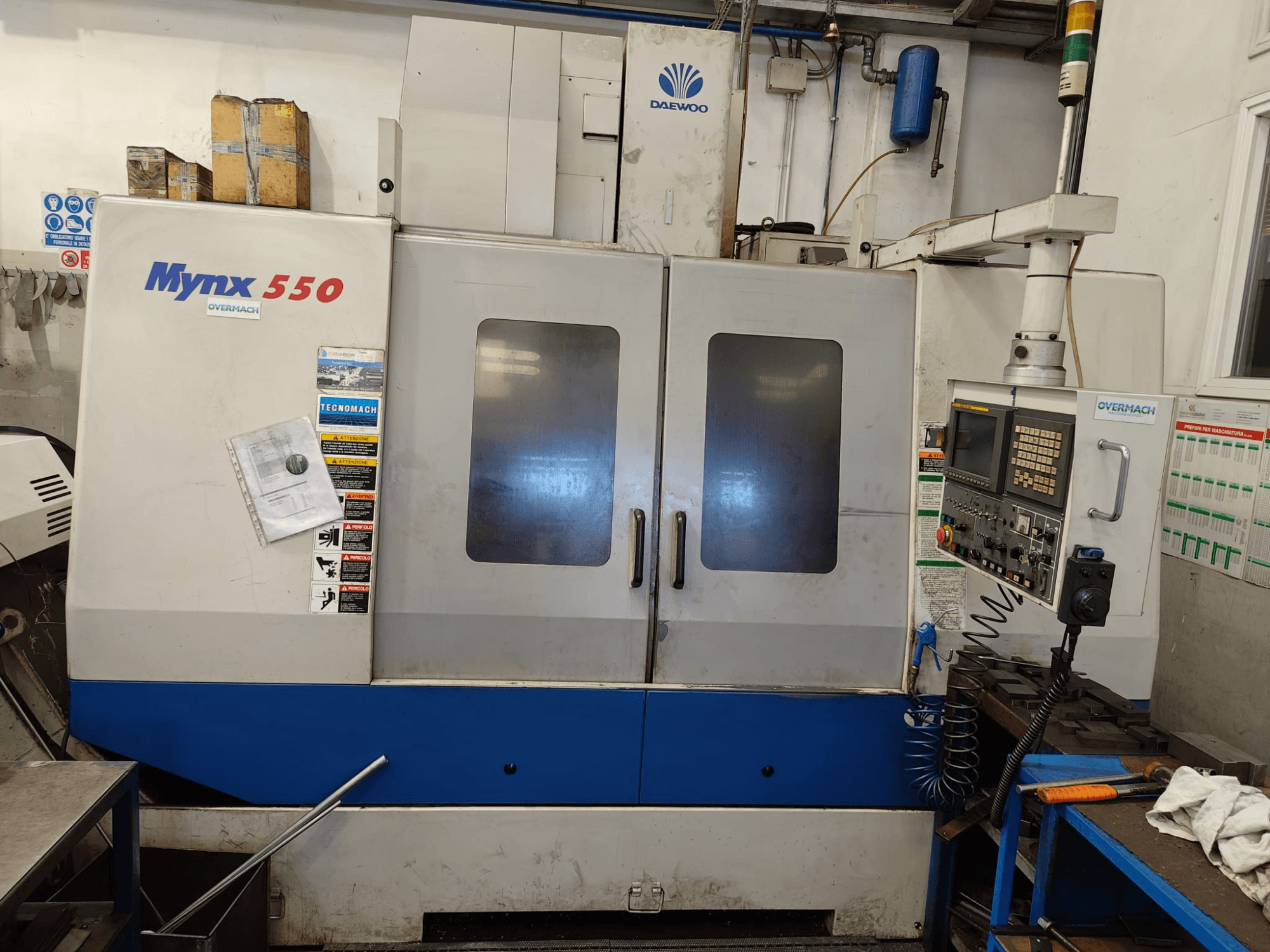 DAEWOO MYNX 550 dikey işleme merkezi, önden görünüm; kontrol paneli, iki büyük kapı ve güvenlik etiketleri ile birlikte.