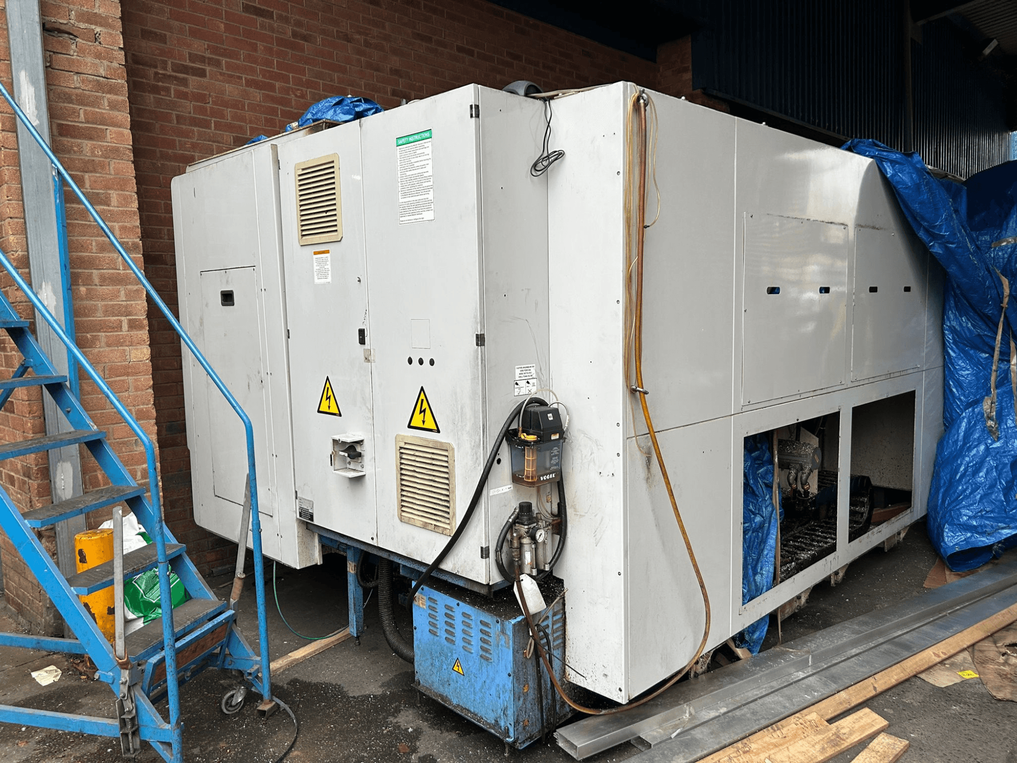 Markası ve modeli belli olmayan büyük bir endüstriyel soğutucunun elektrik bağlantılarını, uyarı işaretlerini ve mavi muşamba örtüyü gösteren yandan görünümü.