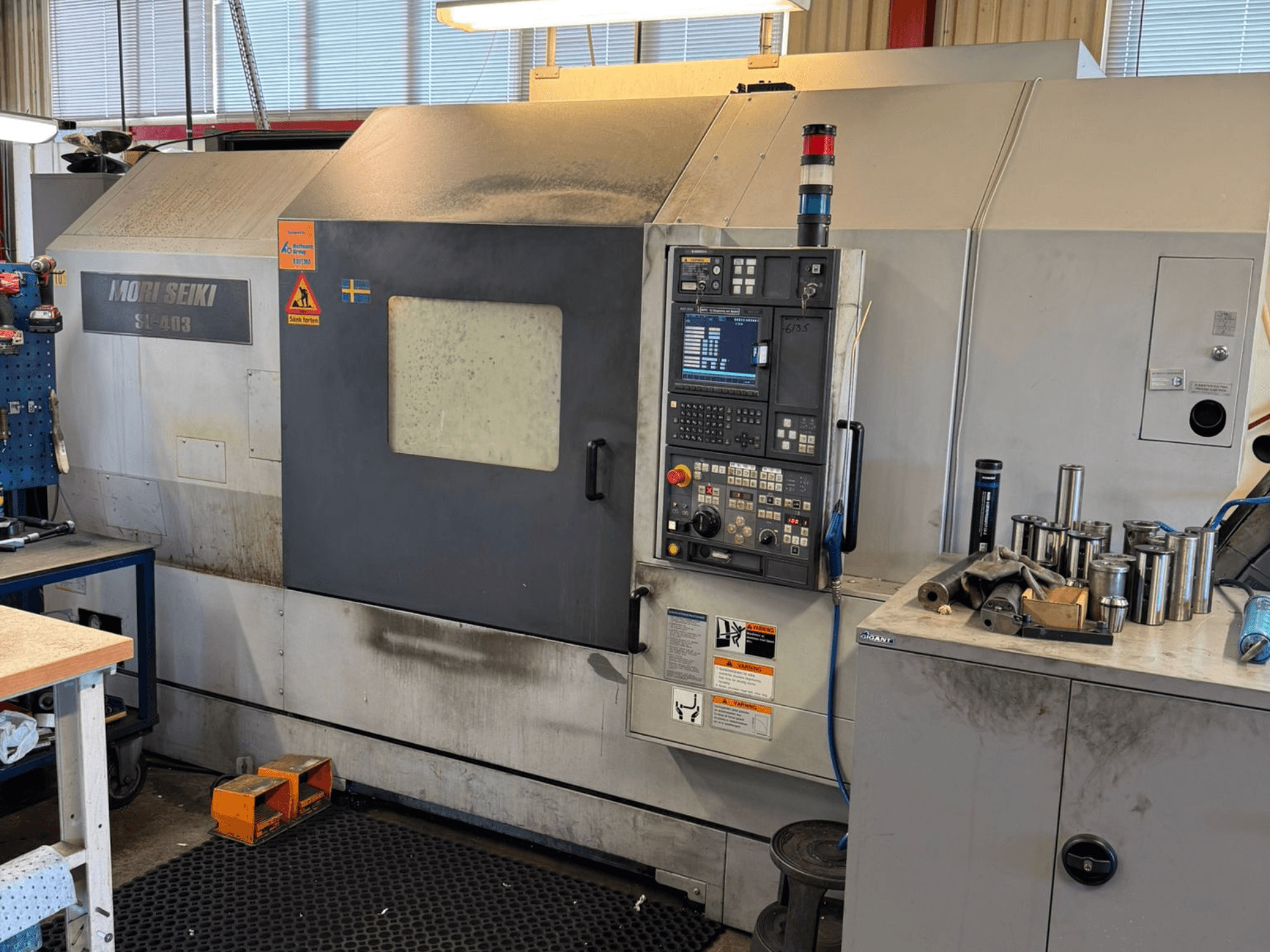 MORI SEIKI SL-403BMC/800 yatay tornalama makinesi atölyede, kontrol paneli ve yakındaki aletleri gösterir, yandan görünüm.