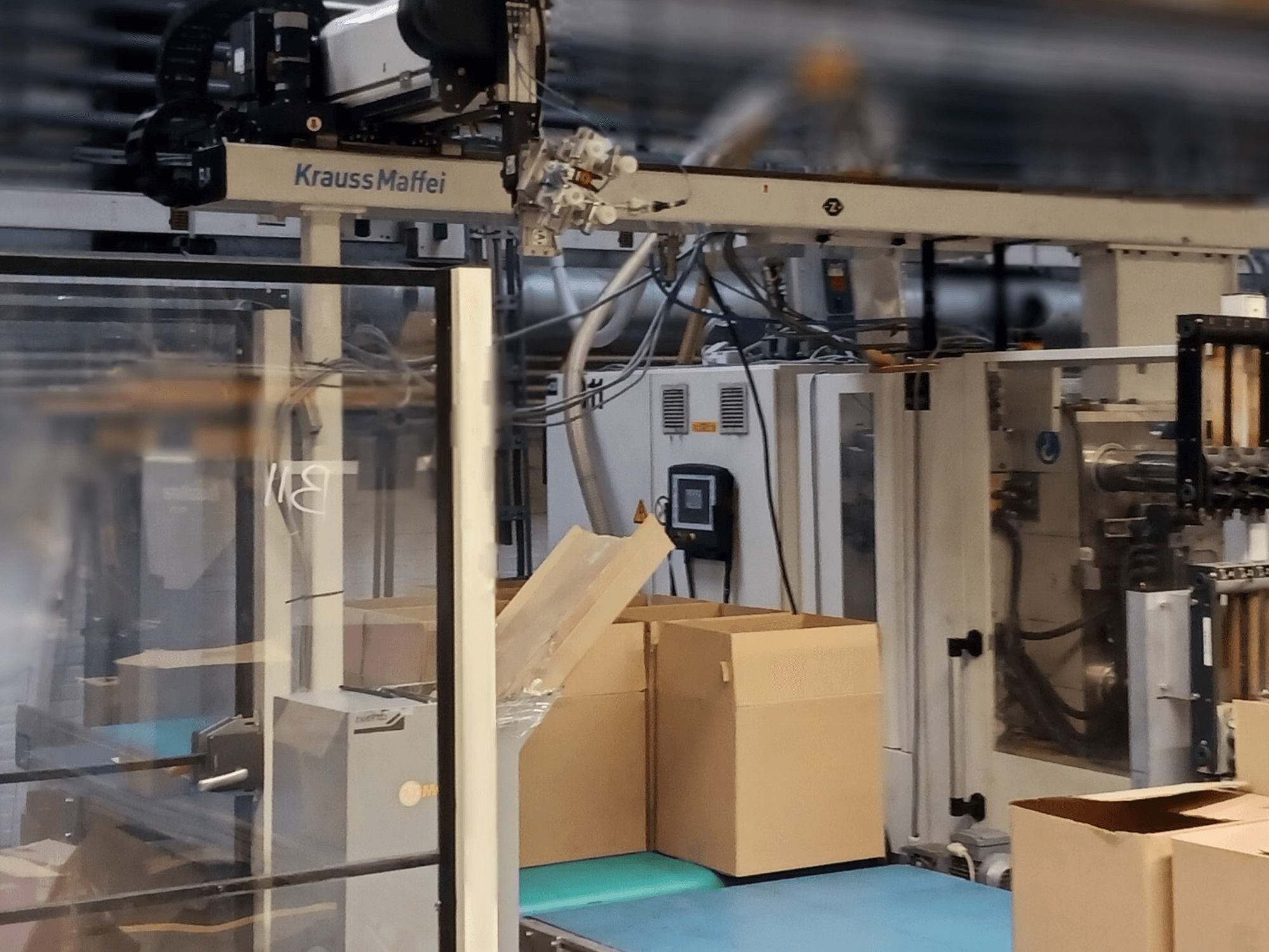Bir fabrikadaki Krauss Maffei otomasyon sistemi, konveyör üzerindeki kutuları ve çalışan makine bileşenlerini gösteren önden görünüm.