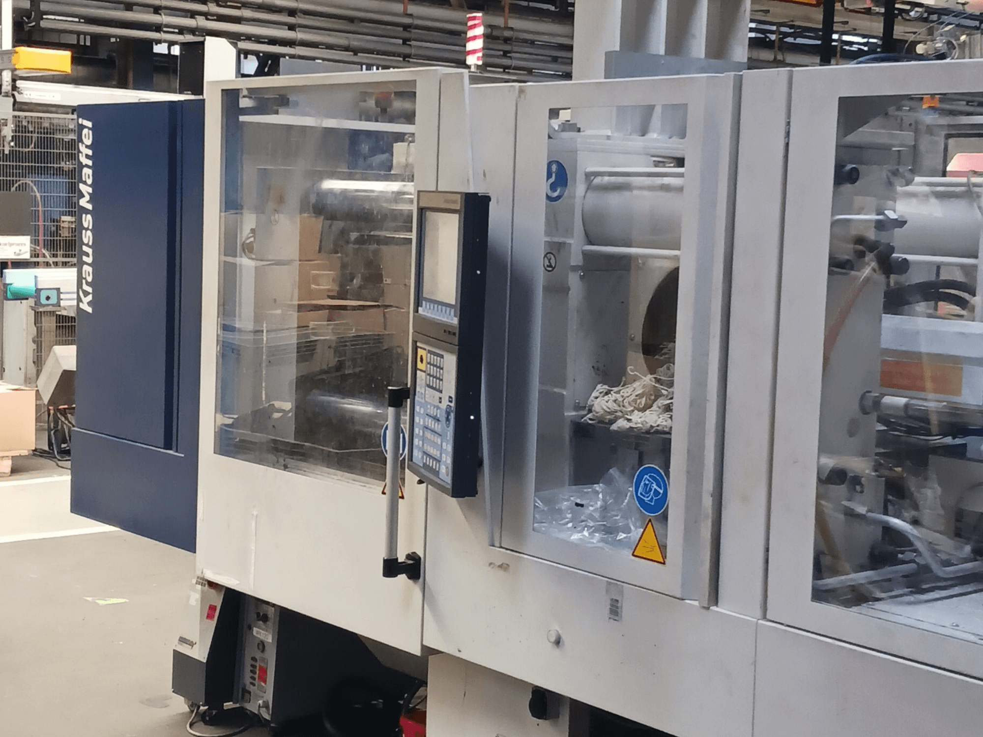 KraussMaffei enjeksiyon kalıplama makinesi, yandan görünüm, arka planda kontrol paneli, makine ve ambalaj malzemelerini gösteriyor.