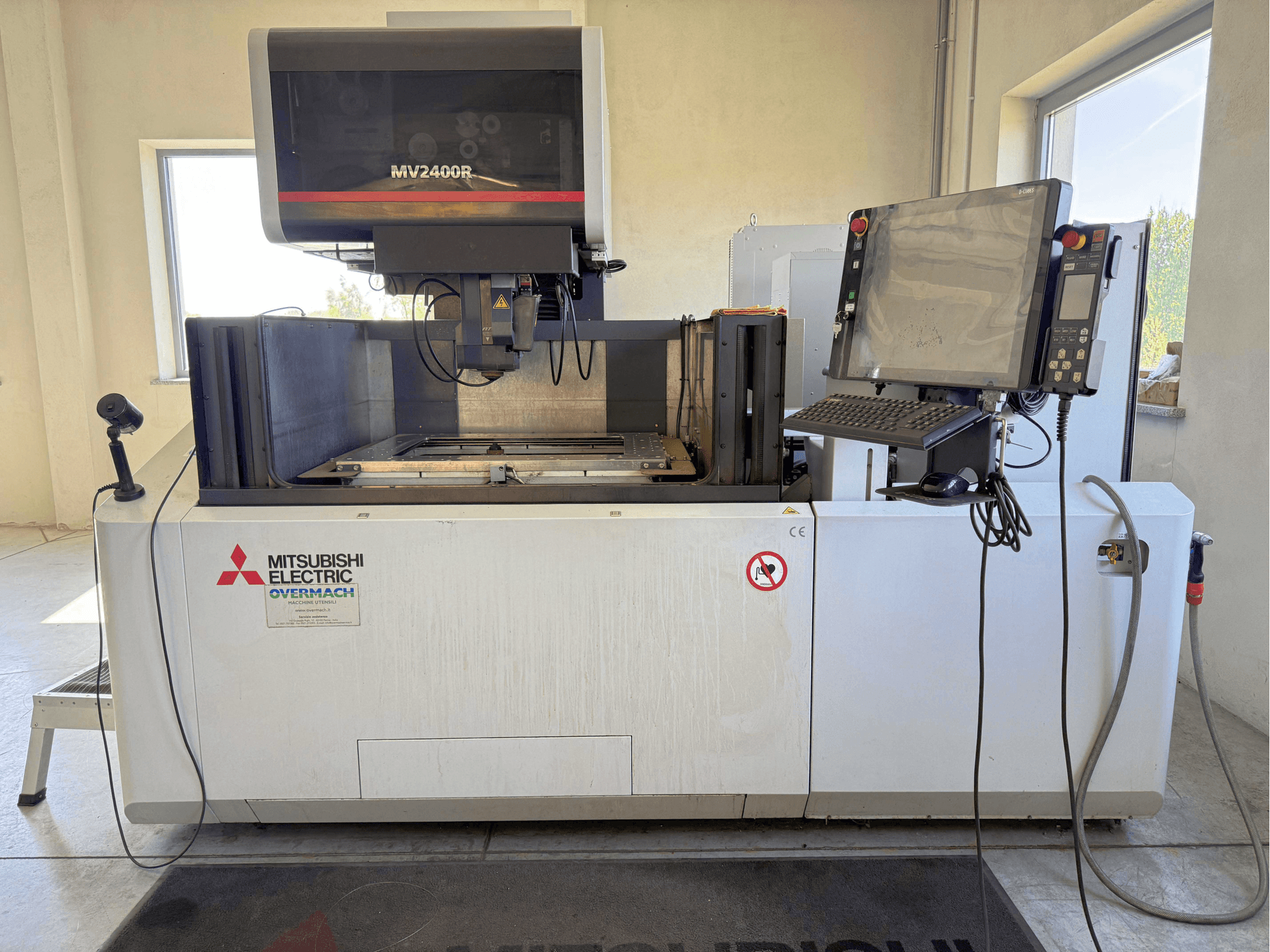 Mitsubishi MV2400R tel EDM makinesinin önden görünümü; kontrol paneli, çalışma alanı ve güvenlik işaretleri görülmektedir.