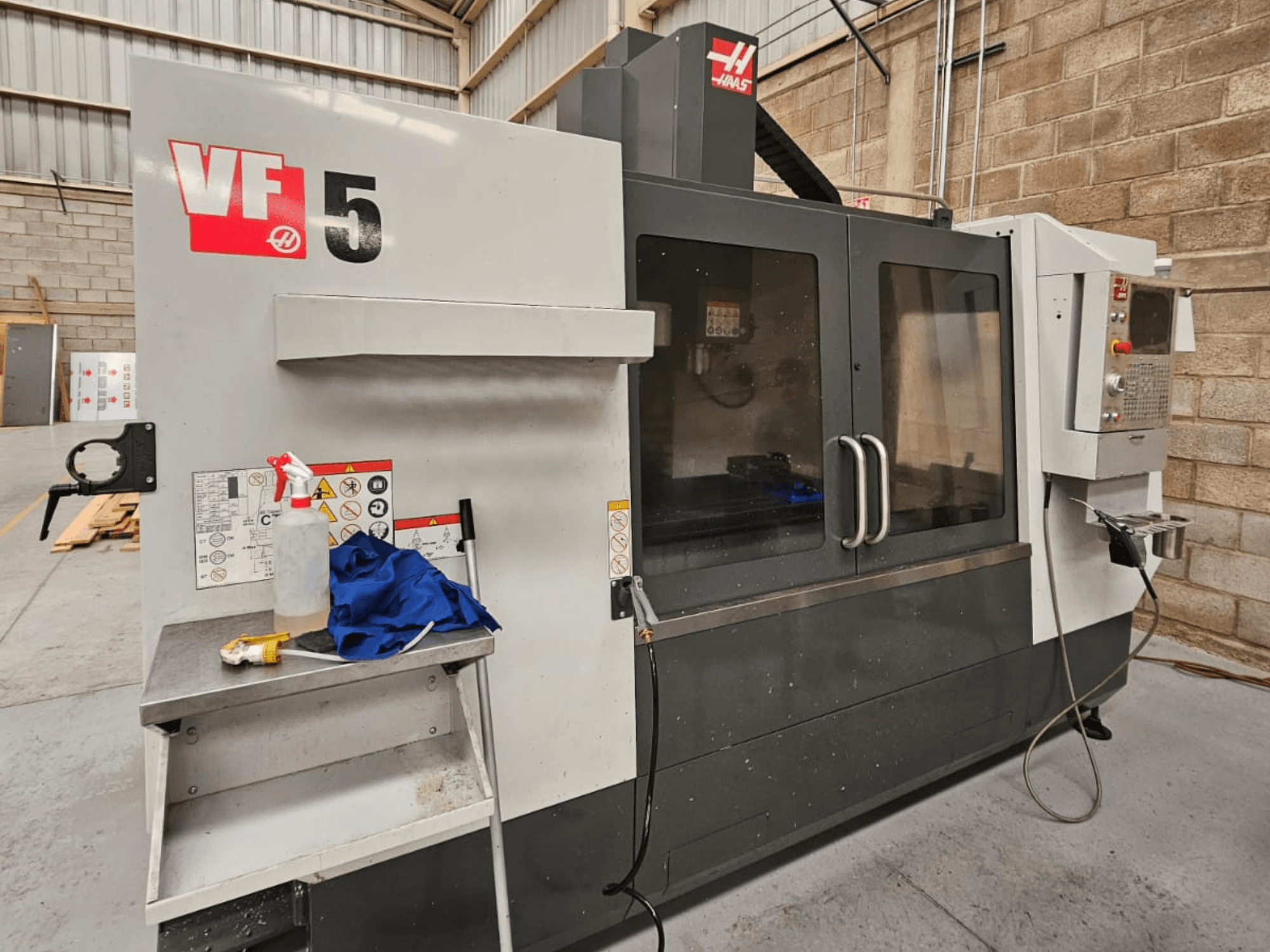 Haas VF-5 CNC freze makinesinin önden görünümü, kontrol paneli, soğutma sıvısı şişesi, mavi bez ve güvenlik talimatları.