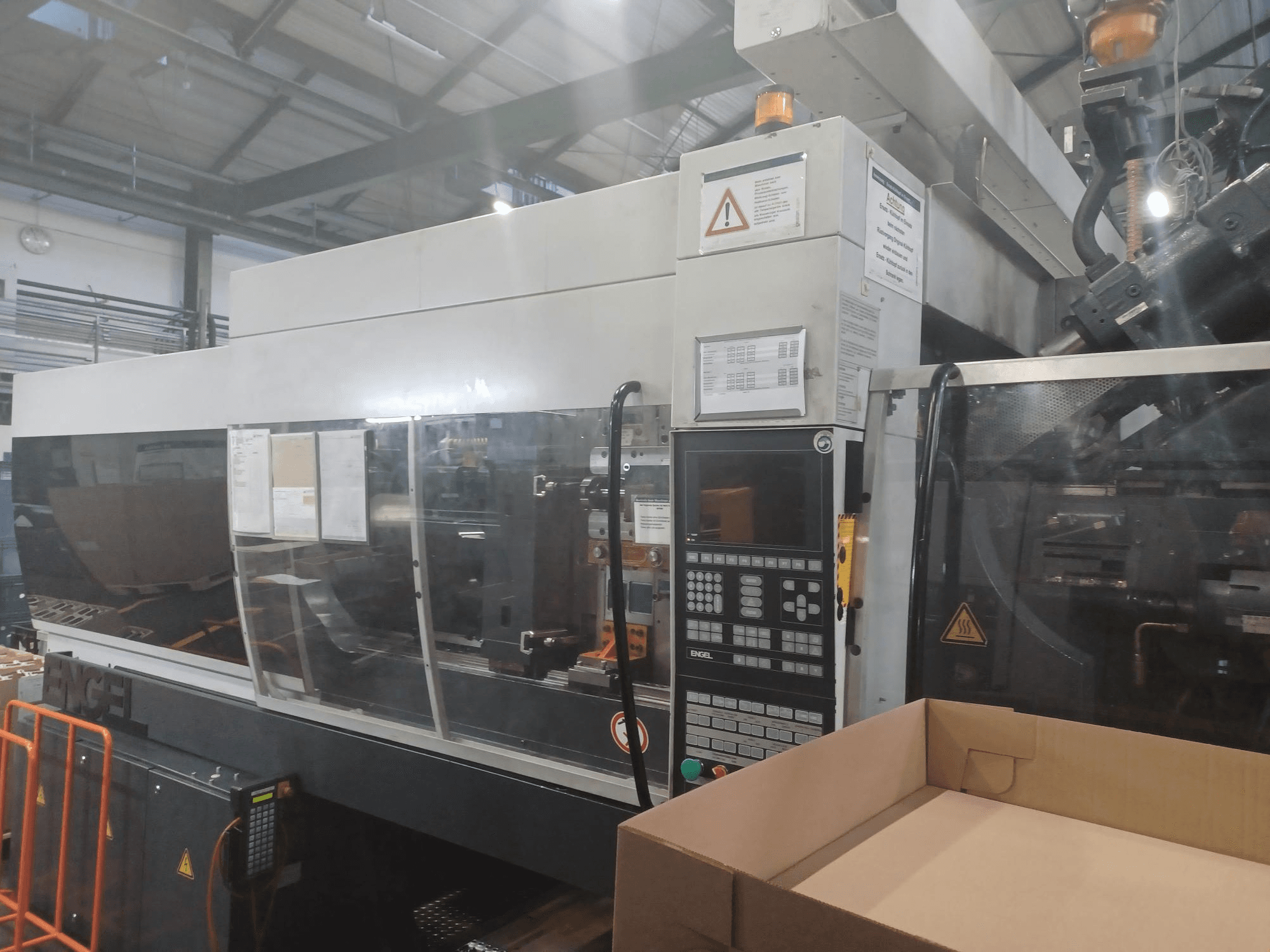 Engel enjeksiyon kalıplama makinesi, önden görünüm, kontrol paneli, güvenlik etiketleri ve yakınına yerleştirilmiş boş bir kutu.