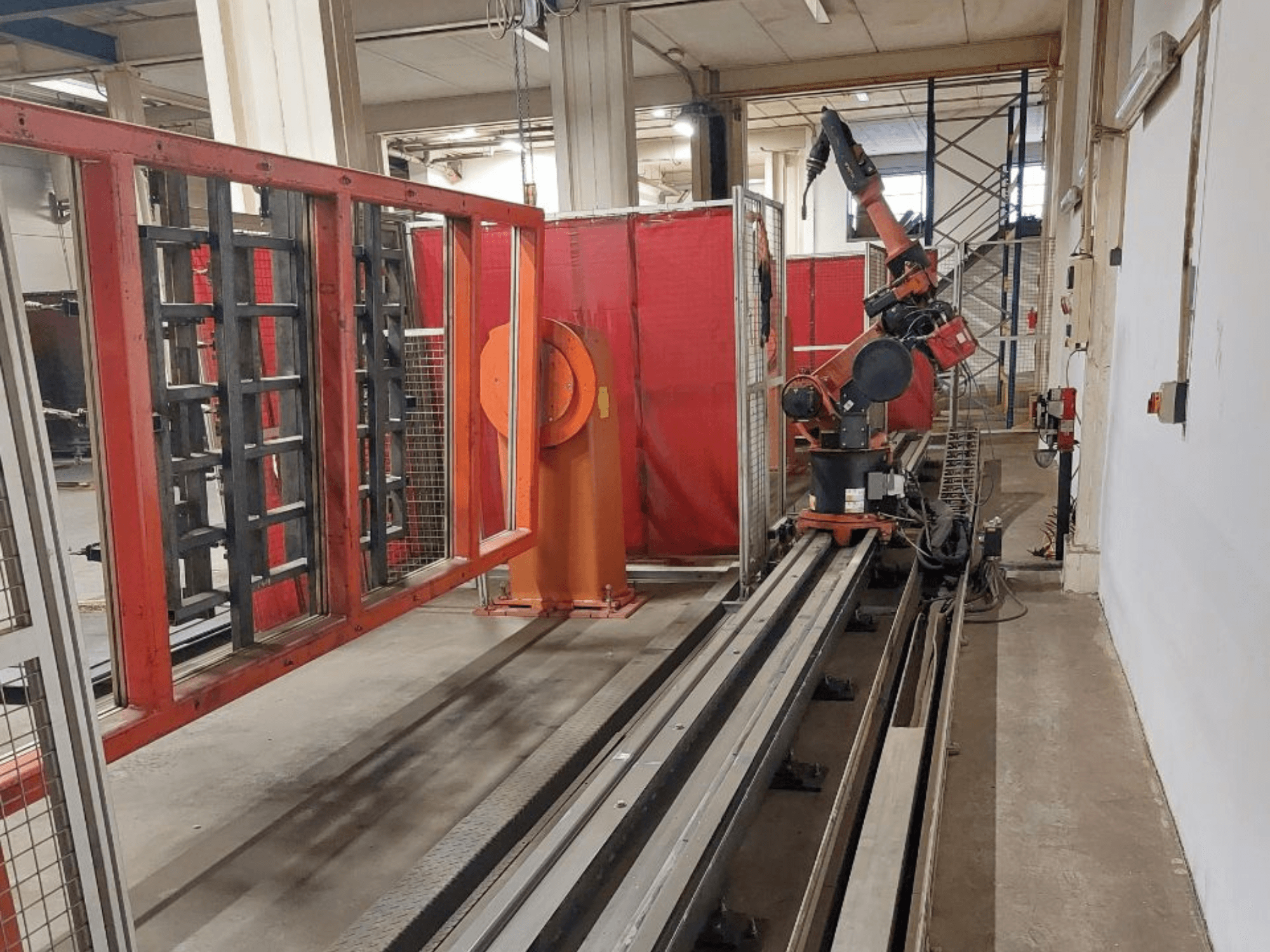 Metal raylar üzerine yerleştirilmiş, kırmızı koruyucu bariyerlerle çevrili, turuncu ve siyah renklere sahip endüstriyel robot kol, önden görünüm.