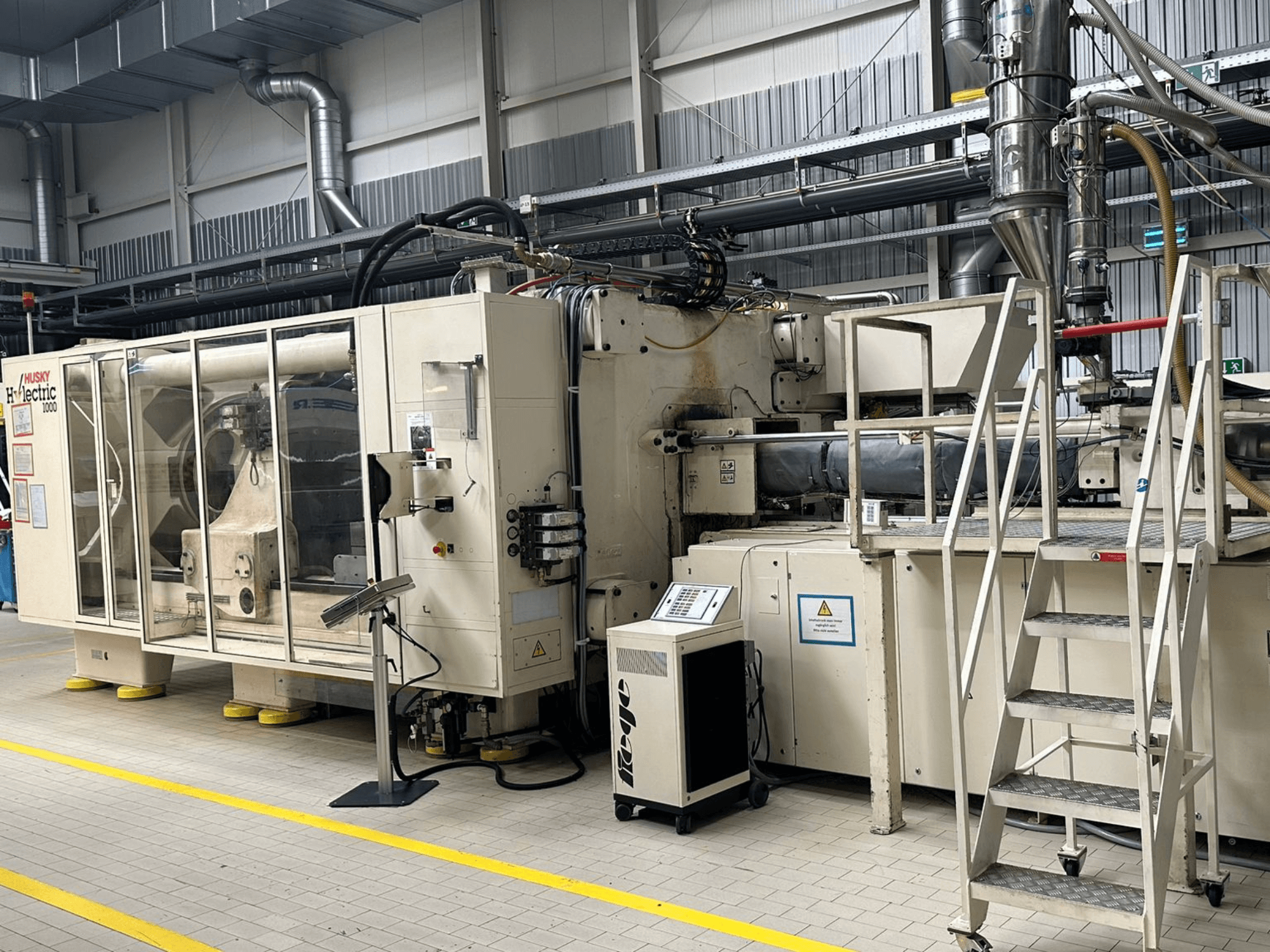 Bir fabrika ortamında çekilmiş, kontrol paneli ve basamakları da görülen Husky Hylelectric H1000 RS95/80 elektrikli enjeksiyon kalıplama makinesinin önden görünümü.