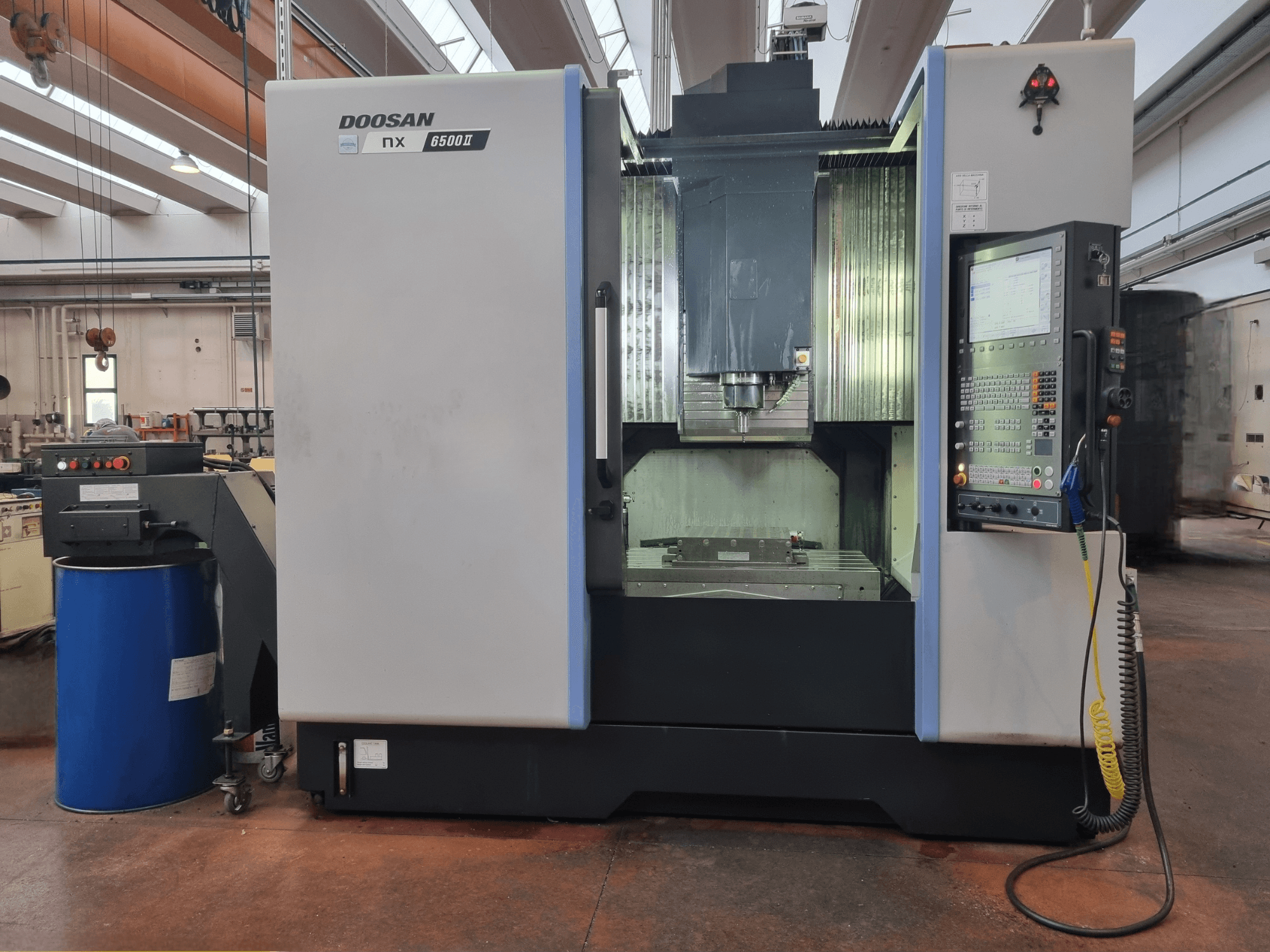 Doosan NX 6500 II CNC makinesi, önden görünüm, kontrol paneli ve işleme alanı, mavi bir varilin yanında konumlandırılmıştır.