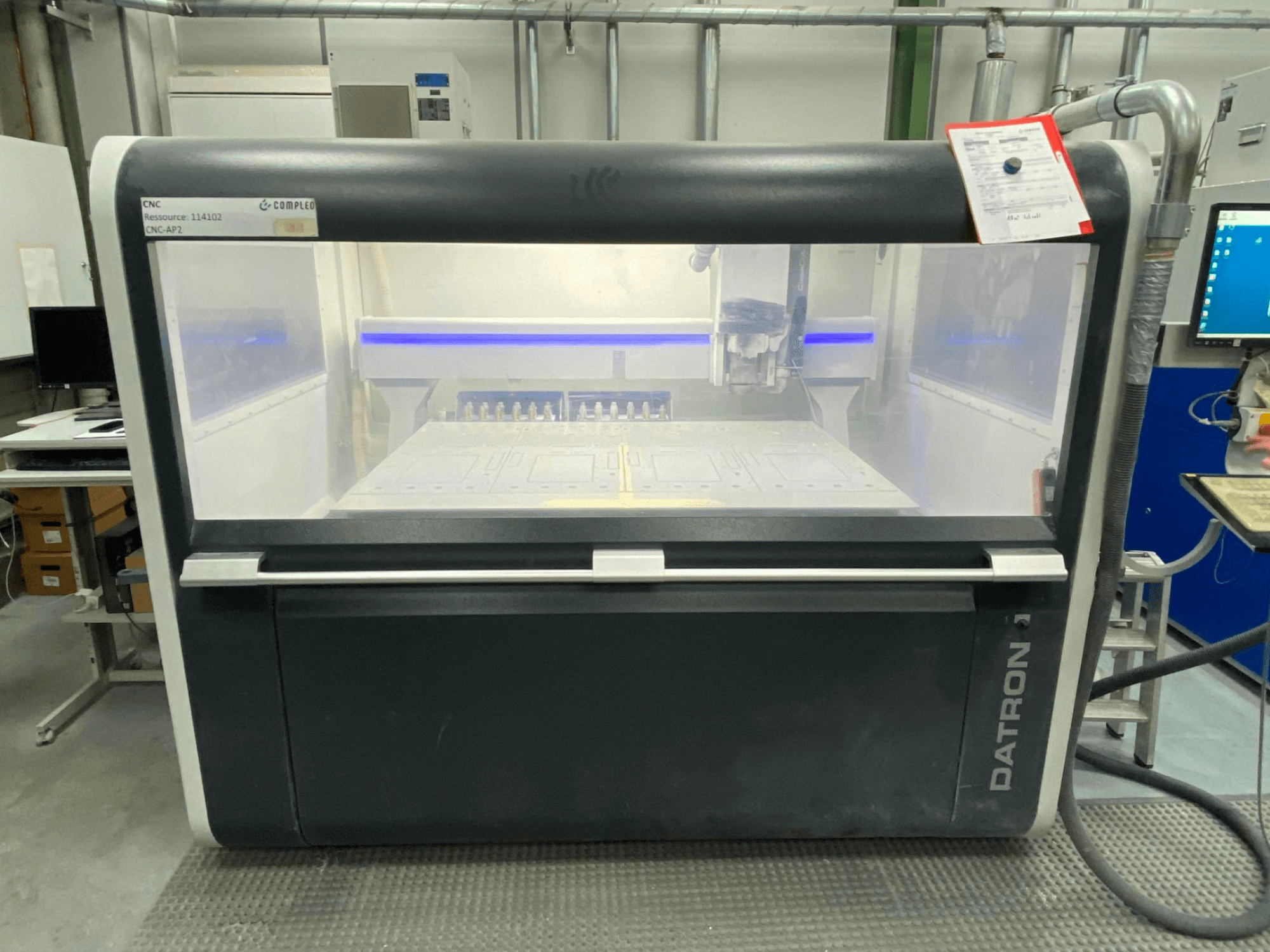 Datron MLCube B dikey işleme merkezinin önden görünümü, görünür kontroller ve bir atölye ortamında bir çalışma masası ile.