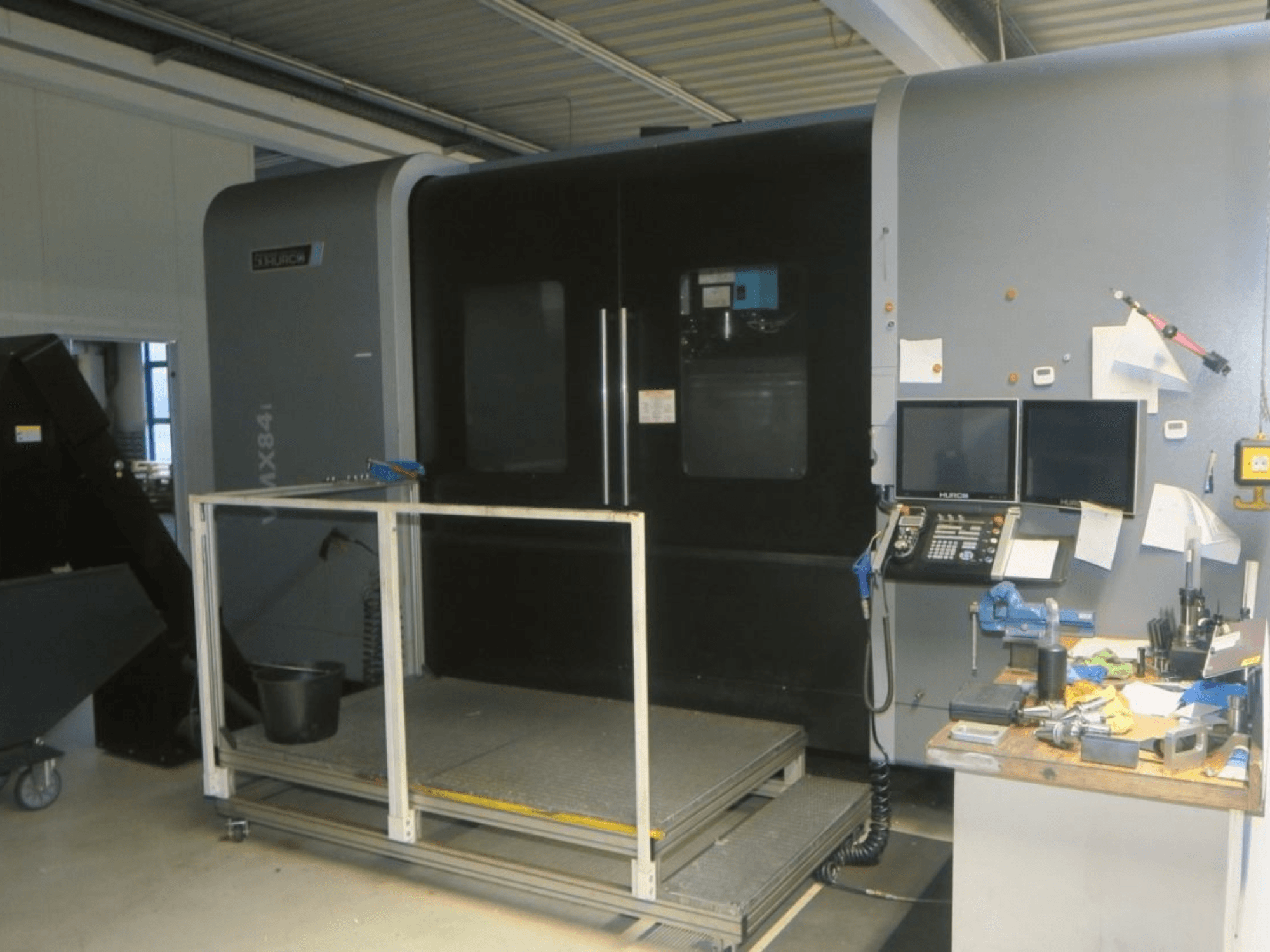 Hurco MX84 CNC makinesinin önden görünümü, çift monitör, kontrol paneli ve çeşitli takımların bulunduğu takım çalışma alanını göstermektedir.