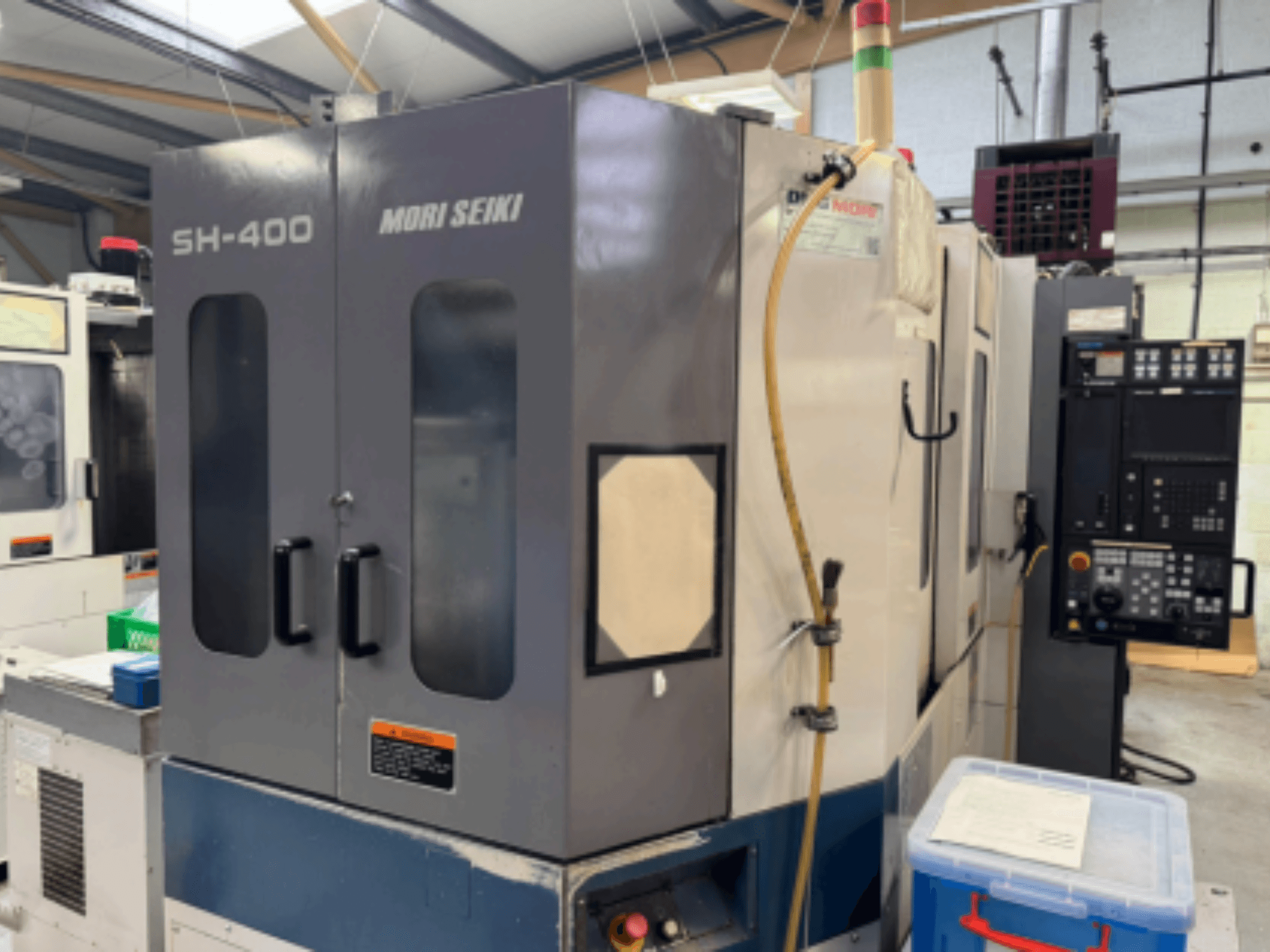 Mori Seiki SH-400 CNC makinesi, önden görünüm, çift kapı, kontrol paneli ve bağlantı hortumu. Mavi saklama kabı yakında.