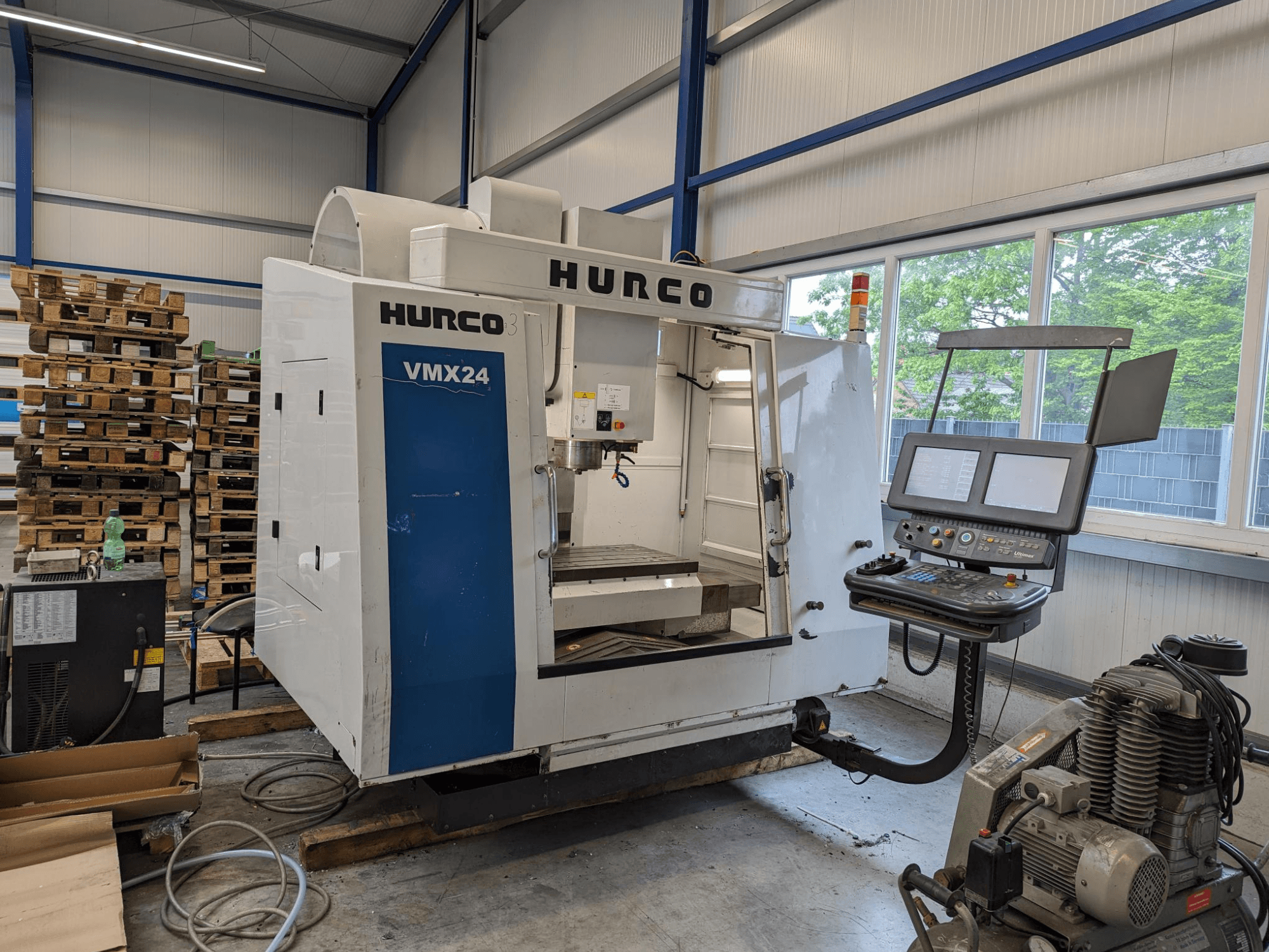 Hurco VMX24 freze makinesi, önden görünüm, kontrol paneli ve takım alanı; arka planda istiflenmiş paletler; endüstriyel çalışma alanı ortamı.