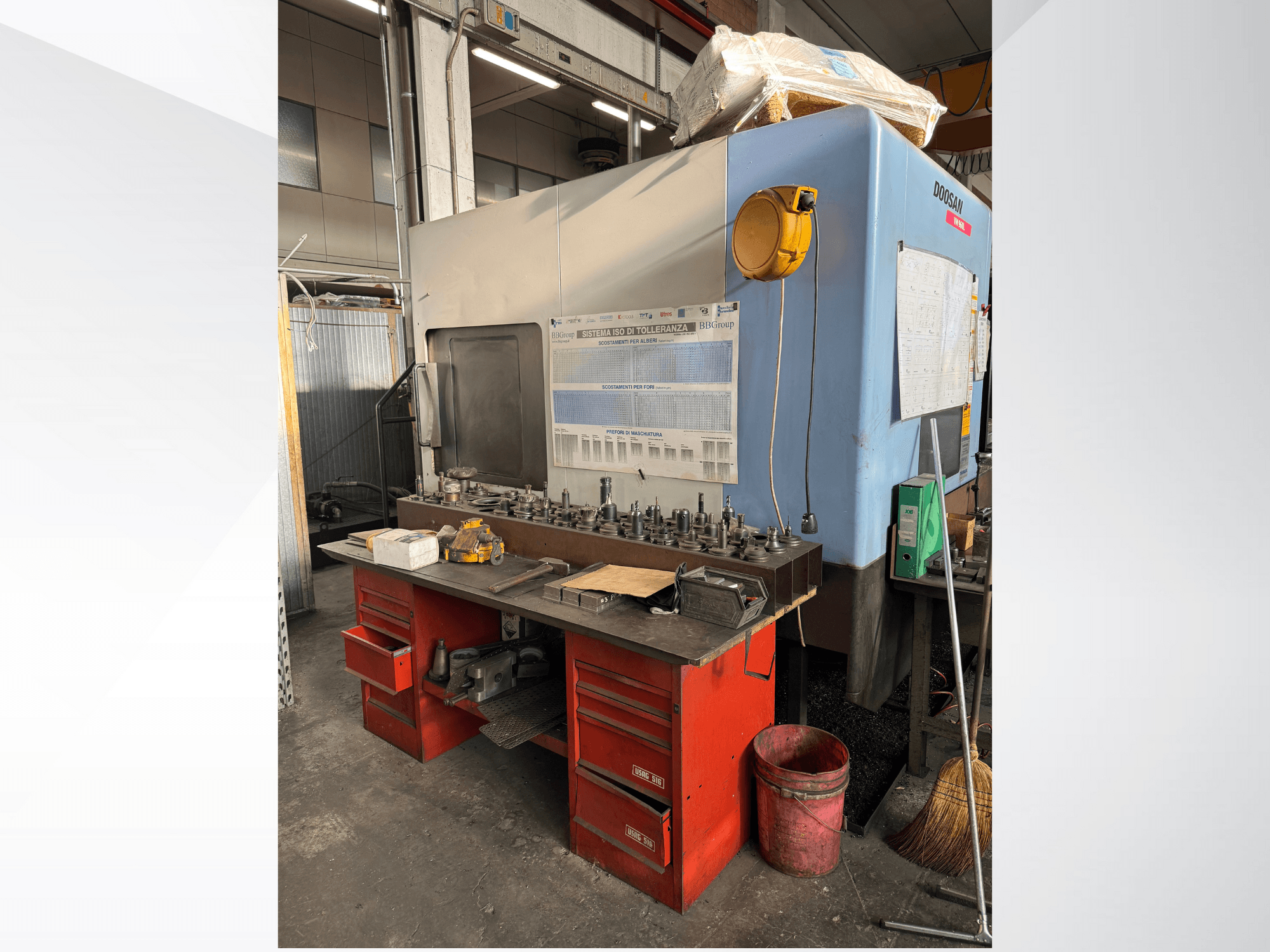 Kırmızı tezgahlı ve depolu, alet ve ekipman sergileyen Doosan işleme merkezi, önden görünüm.