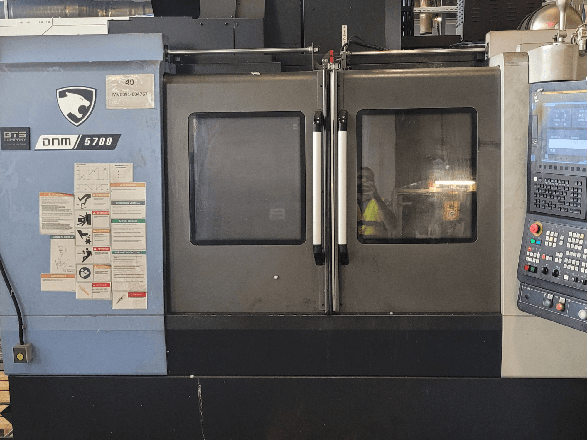DOOSAN DNM 5700 dikey işleme merkezinin, kontrol paneli, güvenlik etiketleri ve hafifçe aralık bırakılmış kapıları ile birlikte önden görünümü.