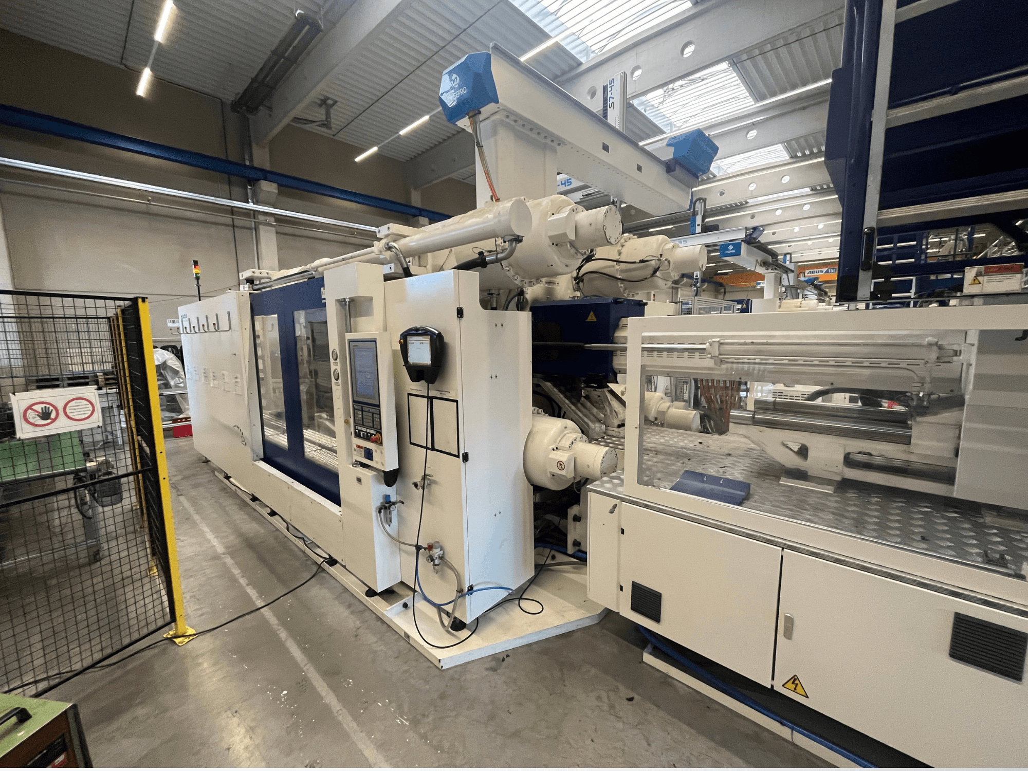 Beyaz ve mavi enjeksiyon kalıplama makinesi, önden görünüm, kontrol paneli, robotik kol ve güvenlik işaretleri.