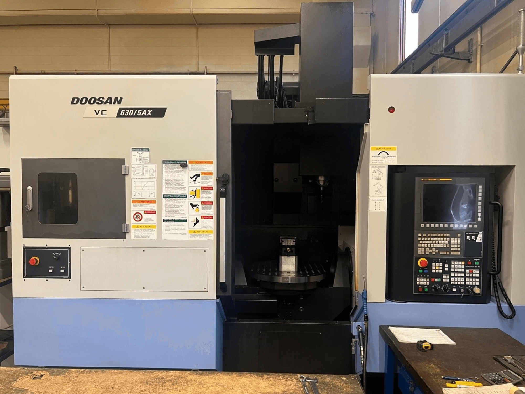 DOOSAN VC 630/5AX dikey işleme merkezinin önden görünümü; kontrol paneli ve iş mili bölümü, üzerinde güvenlik etiketleri açıkça görülebilir şekilde gösterilmiştir.