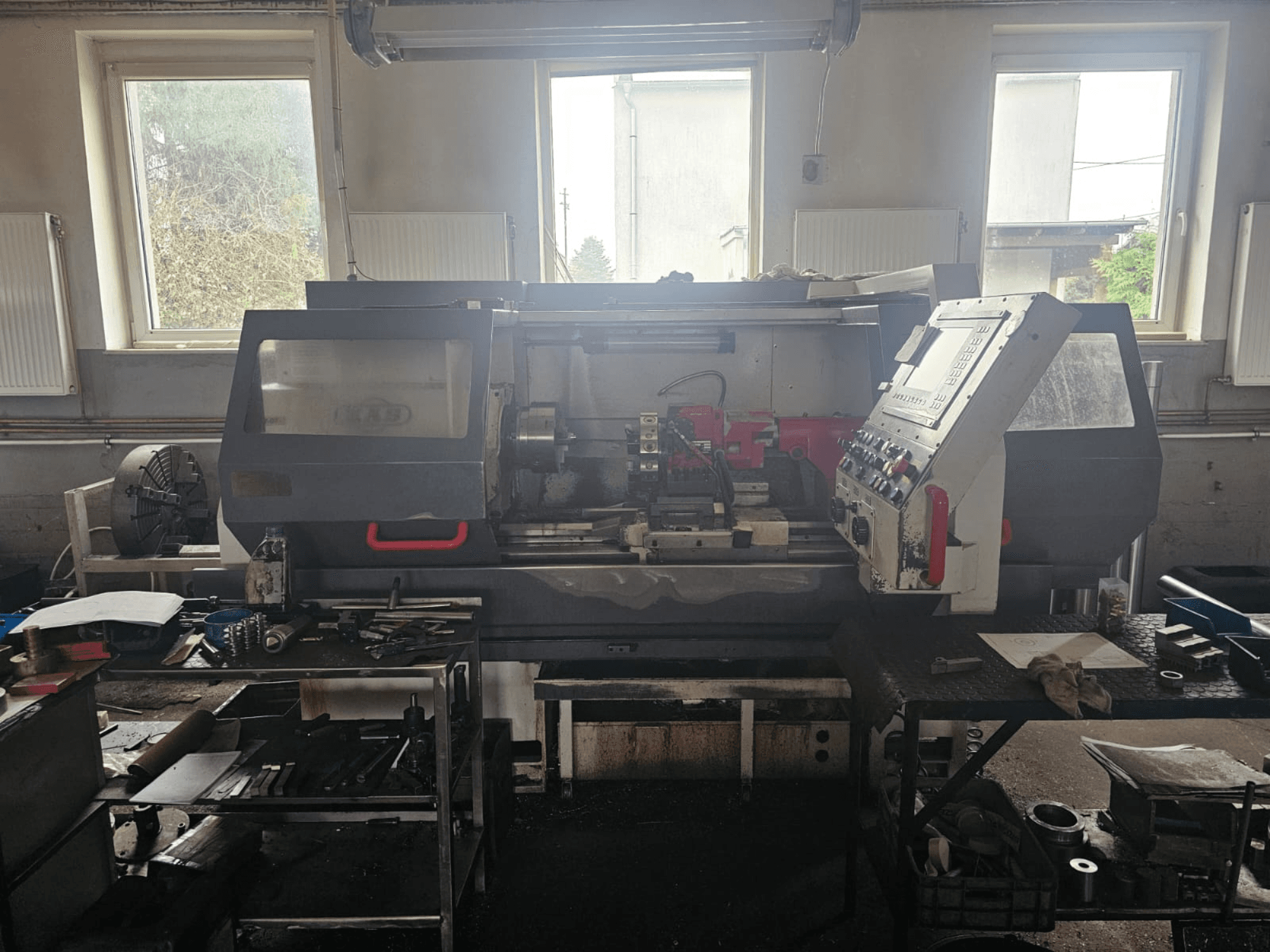 Bir atölyede torna makinesi, önden görünüm, kontrol paneli ve masaların üzerinde aletler, pencerelerle çevrili ve dağınık çalışma alanı.