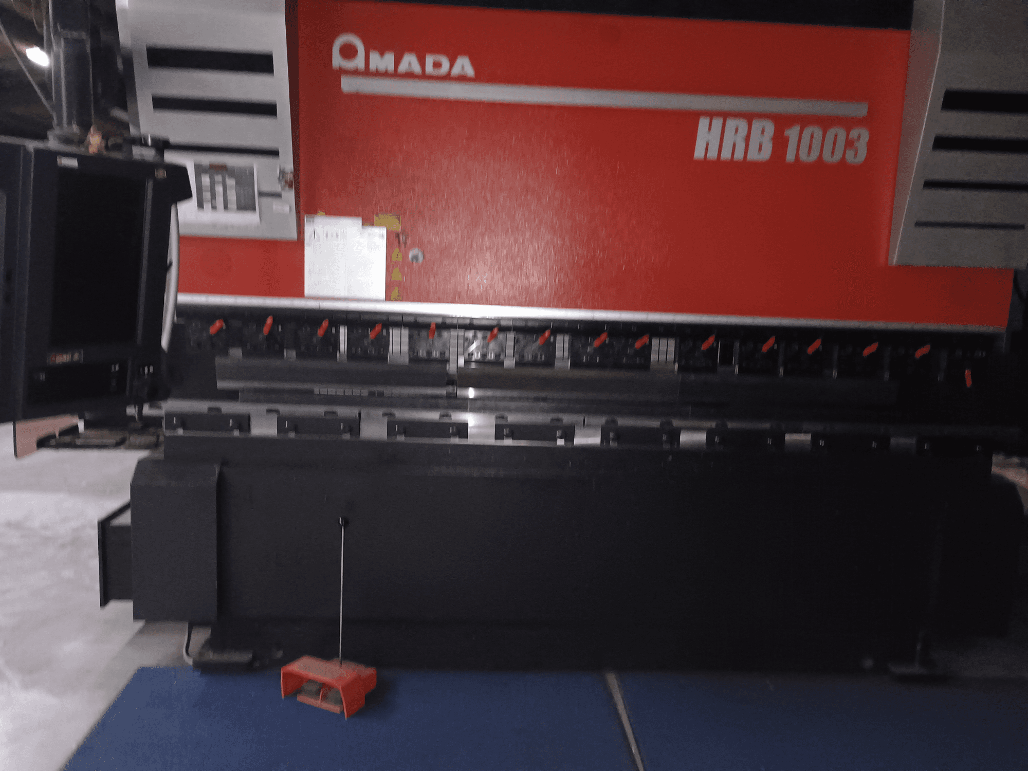 Amada HRB 1003 abkant pres, önden görünüm; kırmızı düğmeli ve siyah kasalı kontrol paneli, endüstriyel bir ortama yerleştirilmiştir.