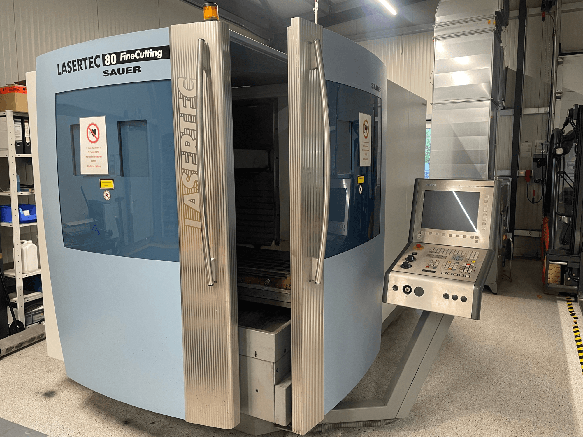 DMG Sauer Lasertec80 FineCutting sac kesme makinesinin önden görünümü; kontrol paneli, açık kapılar ve görünür güvenlik işaretleri ile birlikte.