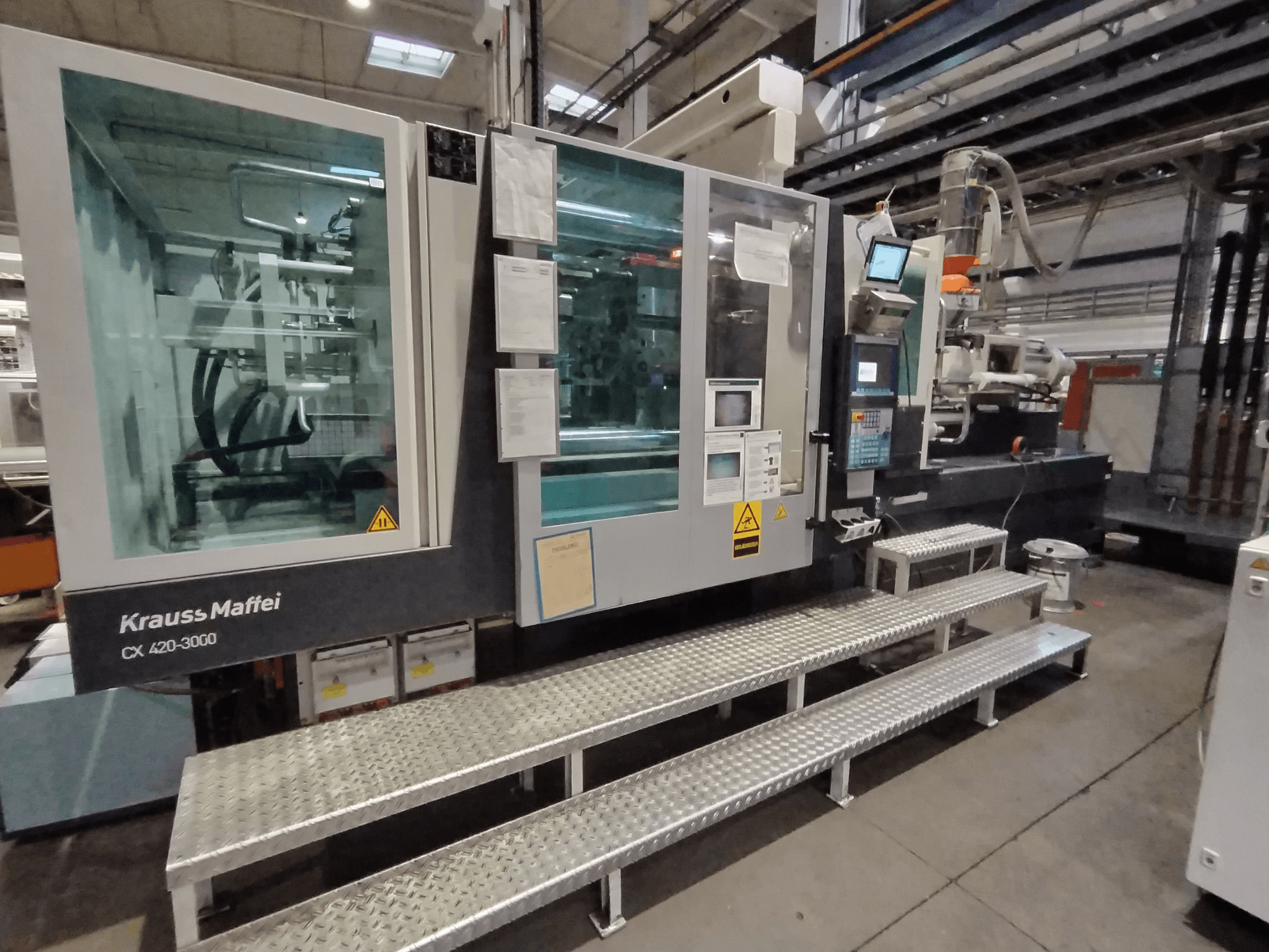 Krauss Maffei CX 420-3000 enjeksiyon kalıplama makinesi, yandan görünüm, kontrol paneli ve güvenlik işaretleri görünür, metal basamaklar üzerine yerleştirilmiş.