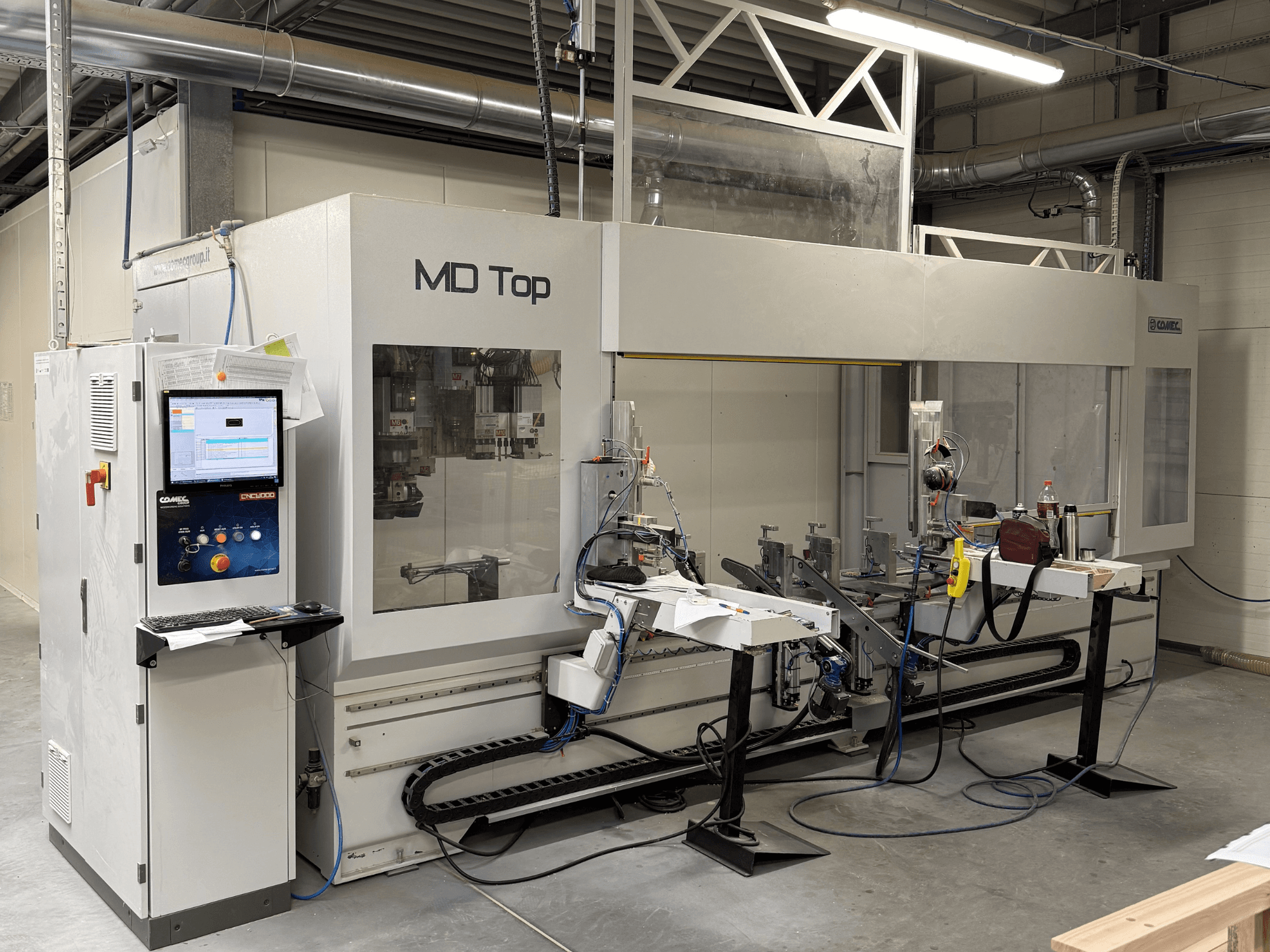 MD Top CNC makinesi önden bakıldığında kontrol paneli, robotik kollar ve alet ve malzemelerin bulunduğu bir çalışma alanına sahiptir.