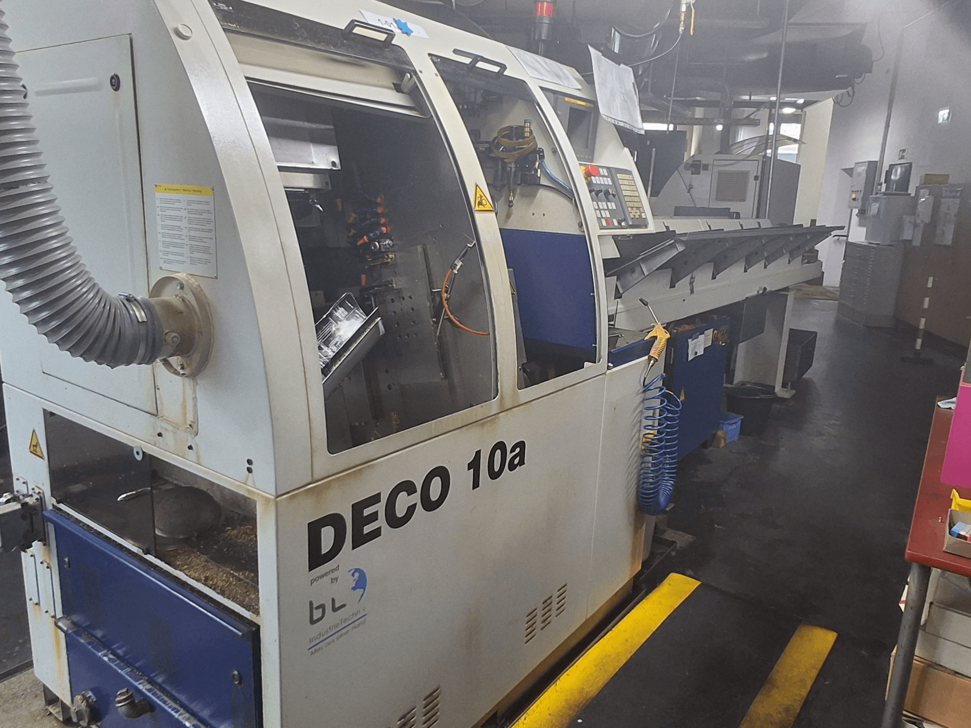 İsviçre Tipi Torna Makinesi TORNOS DECO 10a, önden görünüm; bir kontrol paneli, görünür mekanizmalar ve bir toz emme borusu içerir.