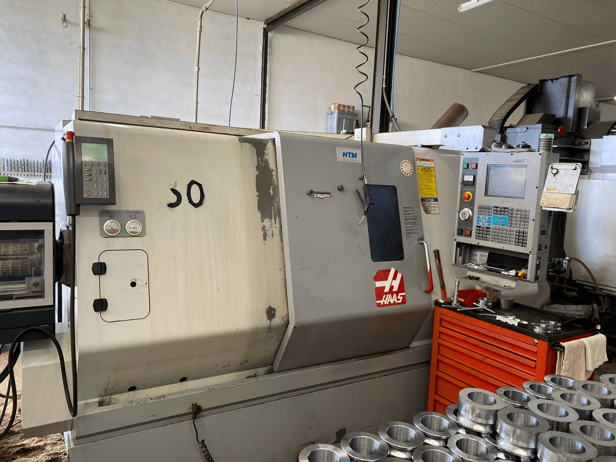 Kontrol panelli Haas CNC makinesi, önden görünüm, işlem düğmelerini, göstergeleri ve yakınlarda istiflenmiş metal parçaları gösteriyor.