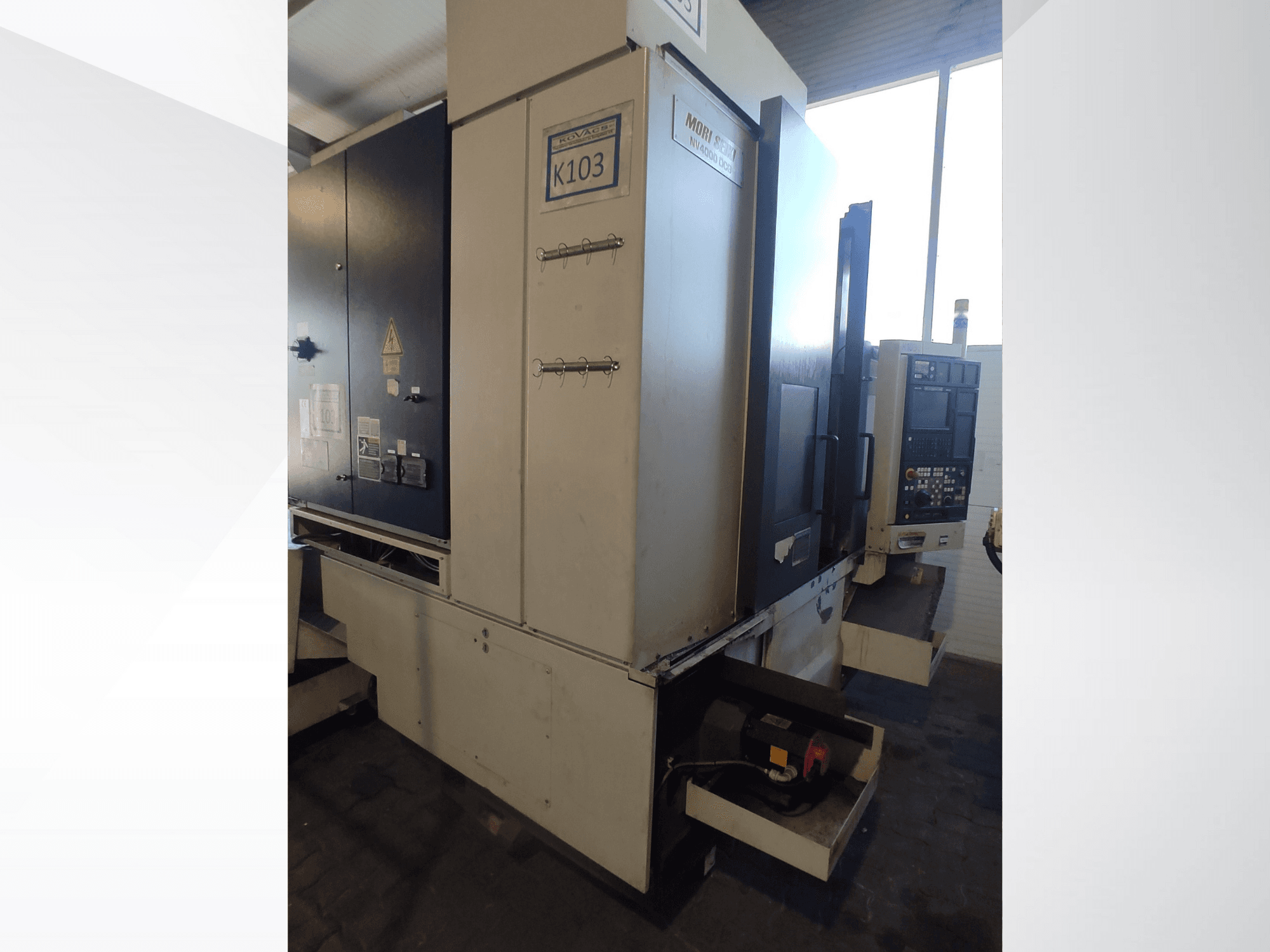Mori Seiki K103 CNC makinesi önden görünümde, kontrol paneli, güvenlik etiketleri ve elektrikli bileşenler görülebilir.
