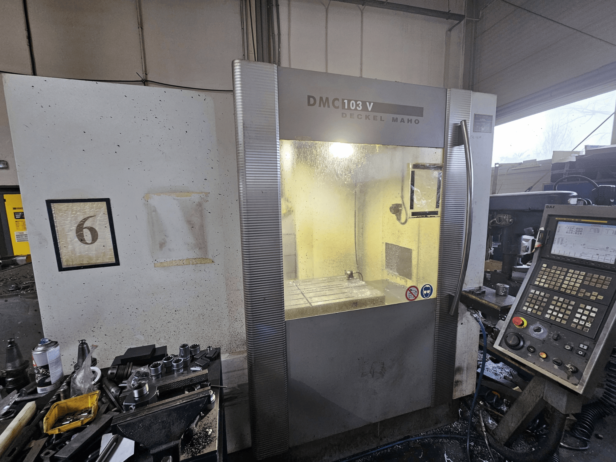 Deckel Maho DMC 103 V CNC makinesi, önden görünüm, kontrol paneli ve çalışma alanı, duvarda '6' rakamı.