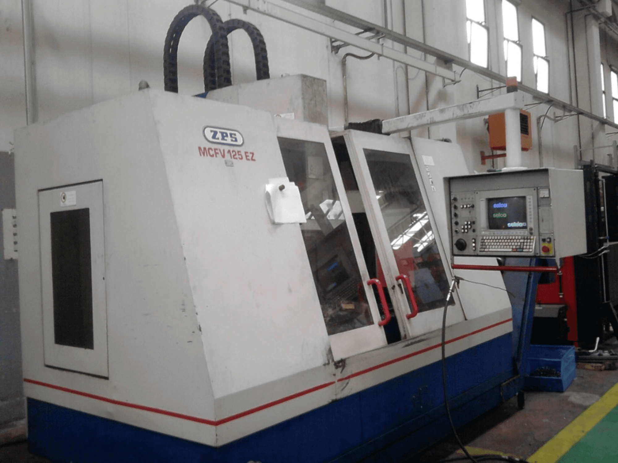 ZPS MCFV 125 EZ CNC makinesi, kontrol paneli ve kırmızı kollarla önden görünüm, görünür hortumlarla bir atölye ortamında.