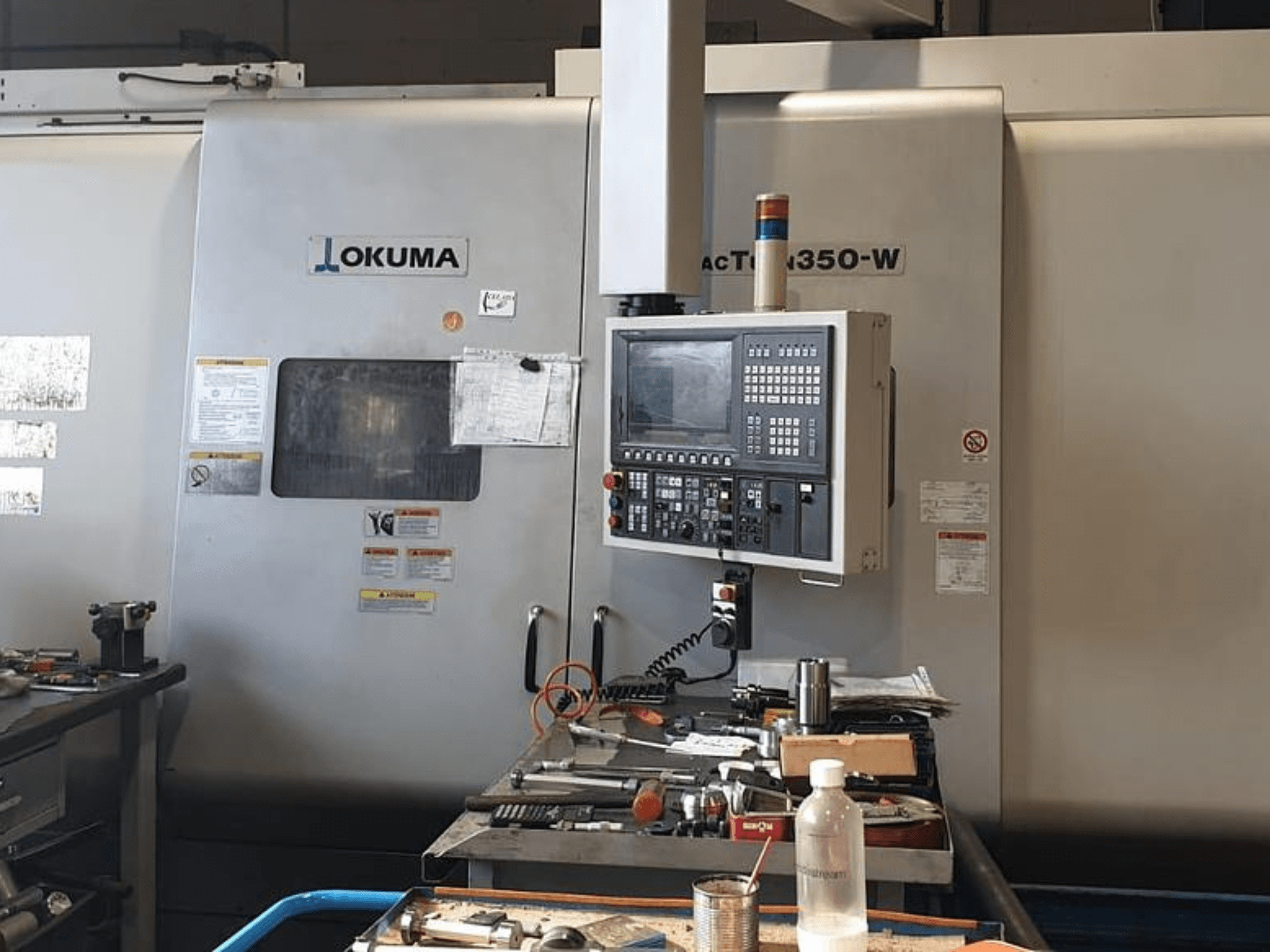 Okuma MAC TURN 350-W torna freze merkezi, kontrol paneli ile önden görünüm ve aletler ve bir şişe ile dolu çalışma alanı.