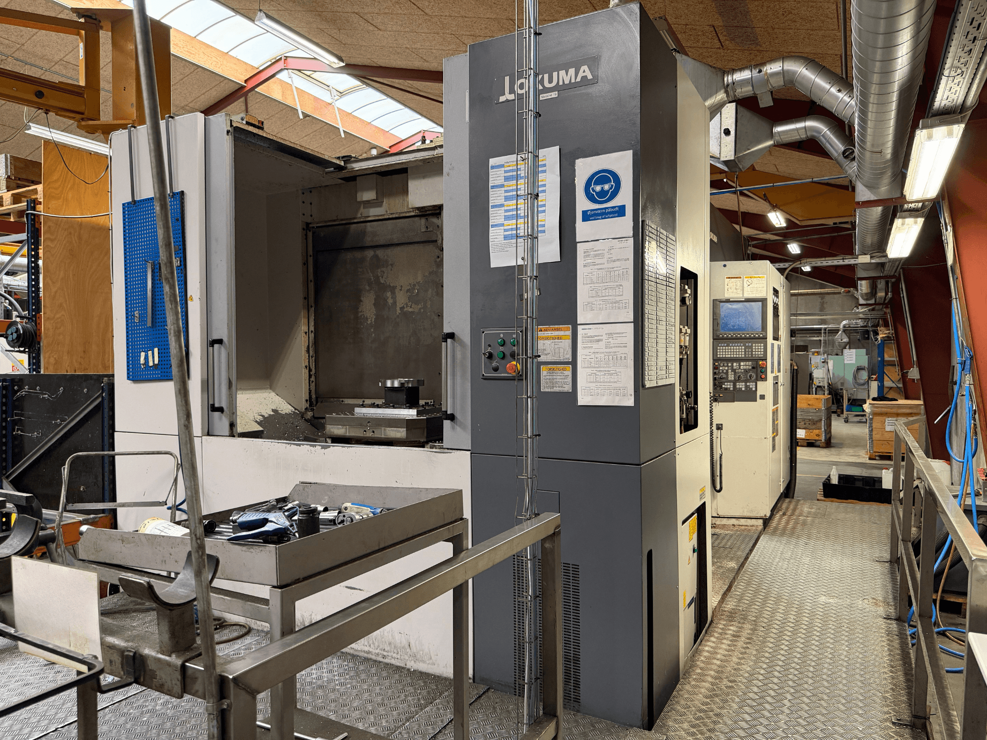 Okuma MA-600HB yatay işleme merkezi, önden görünüm; metal bir tabla üzerindeki takımlar ve makine kumandaları görülmektedir.