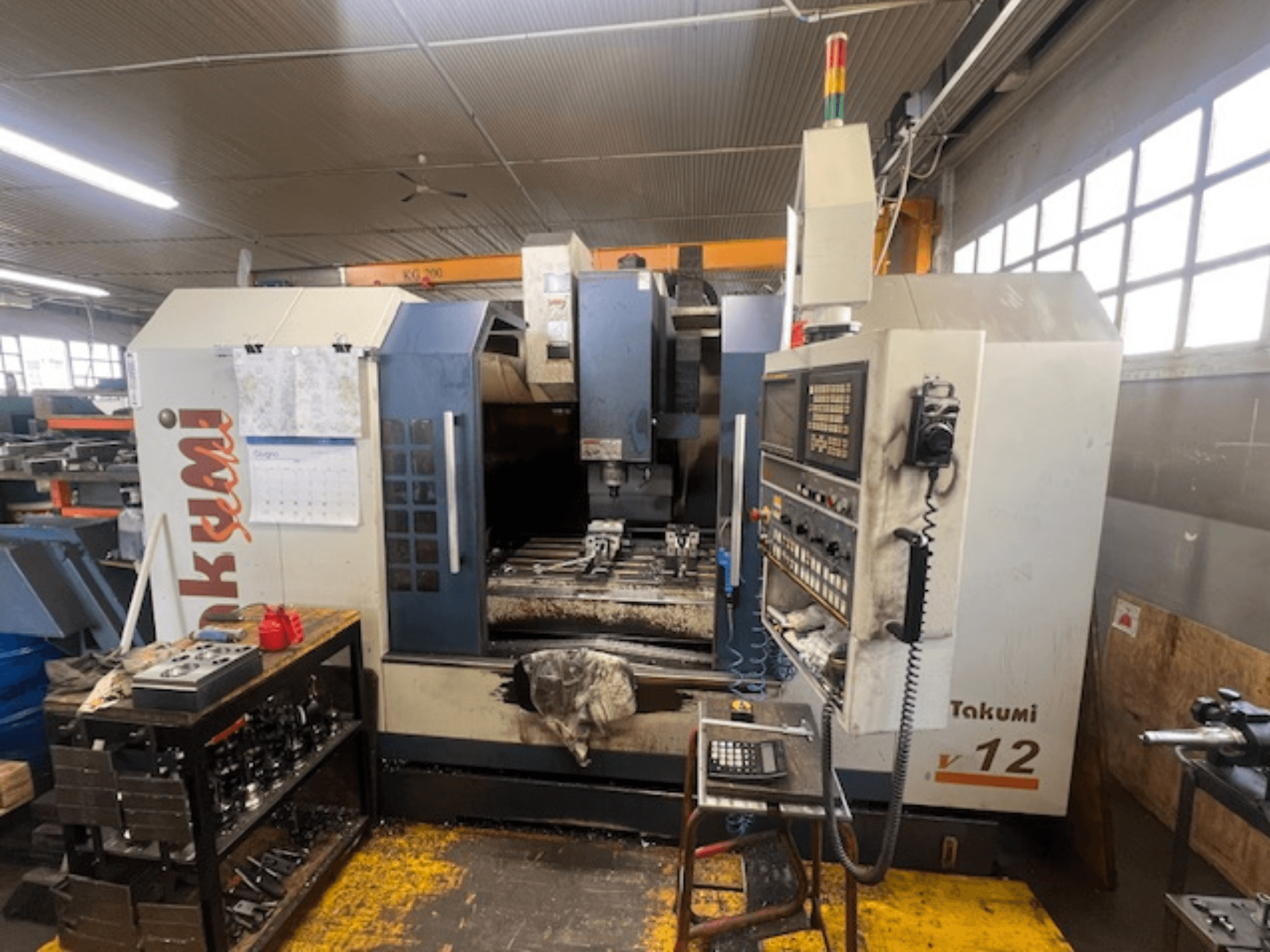 Takumi V12 CNC freze makinesinin yandan görünümü, kontrol panelini, kesim alanını ve iş istasyonundaki takımları gösterir.