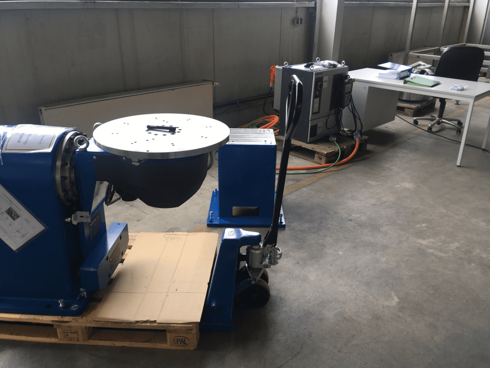 Ofis ortamında ahşap bir palet üzerinde, önden bakıldığında dairesel bir tabana ve kontrol paneline sahip mavi endüstriyel ekipman.