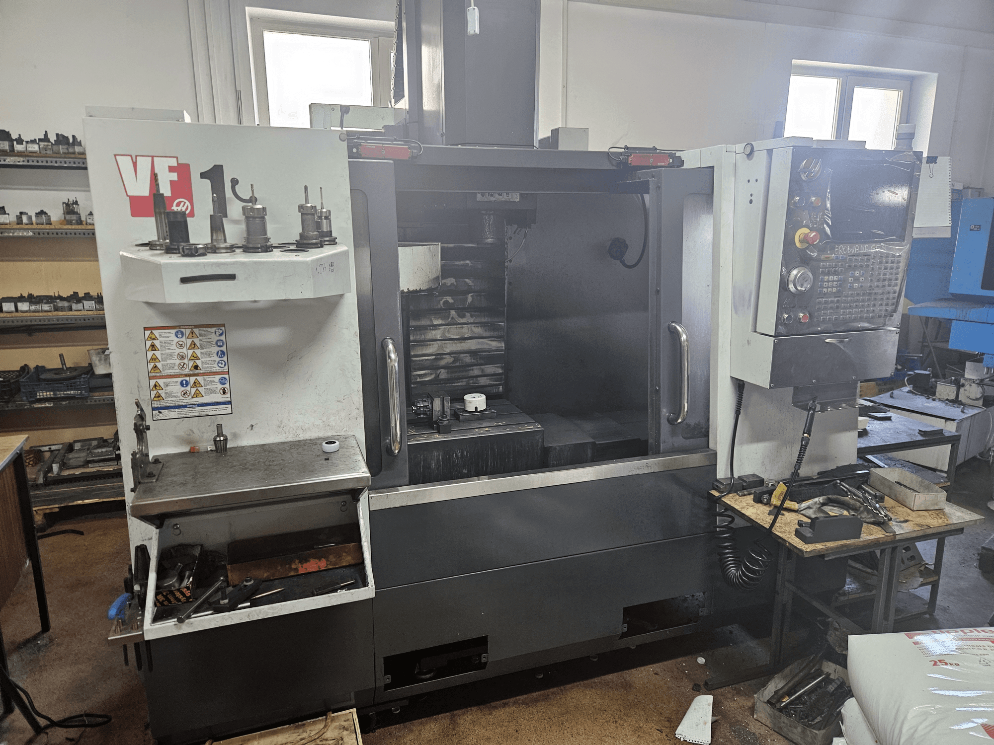 HAAS VF-1 dikey işleme merkezi, takımlar yanda, kontrol paneli görünür ve çalışma alanı arka planda olacak şekilde önden görünüm.