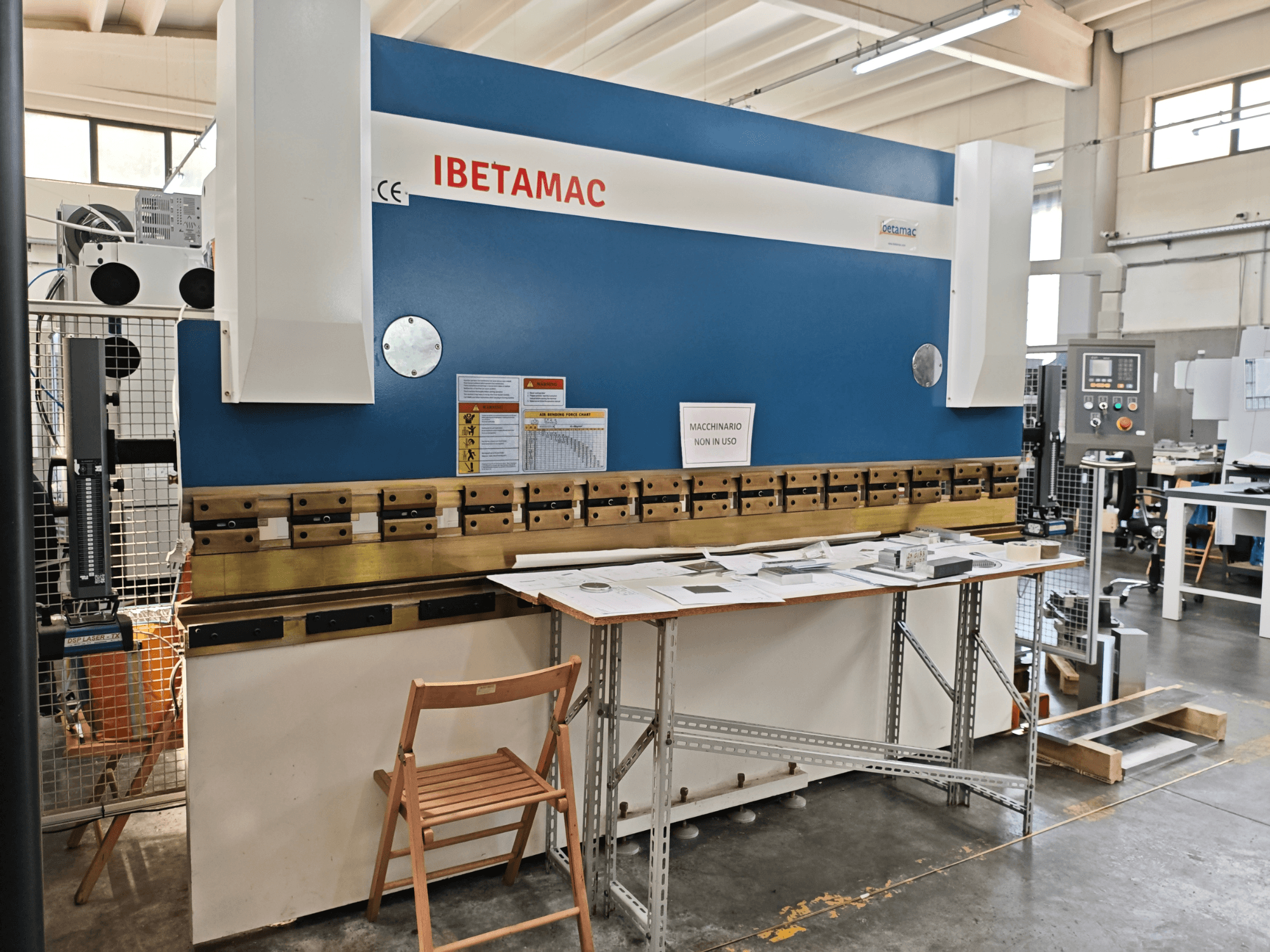 Bir atölyede bulunan IBETAMAC 3200 100T pres fren makinesi; kontrol paneli, çalışma alanı ve güvenlik uyarılarını gösteren yandan görünüm.