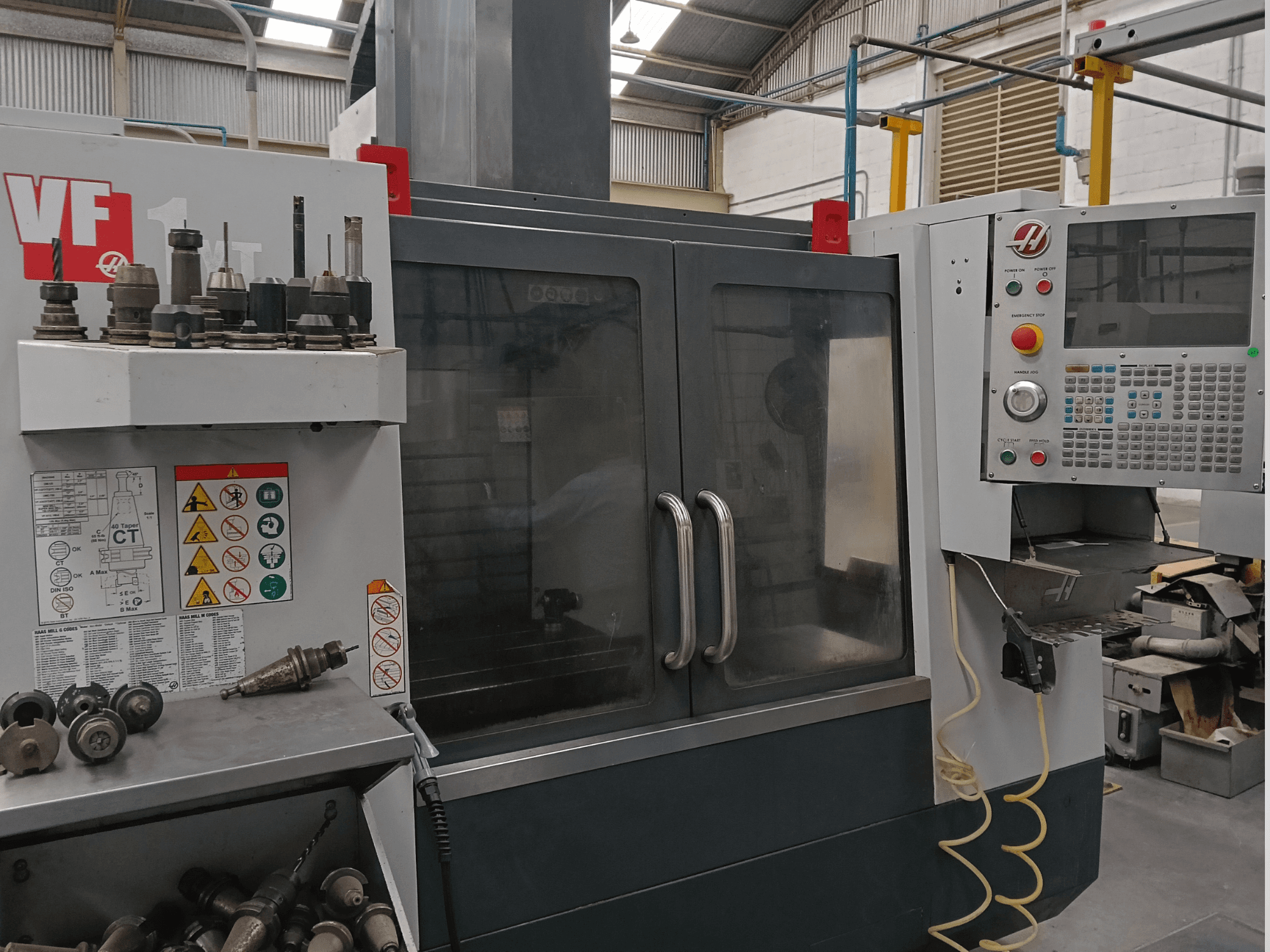 HAAS VF-1YT-3 Dikey İşleme Merkezi’nin önden görünümü; kontrol panelleri, takım tutucular ve görülebilen güvenlik etiketleri gösterilmektedir.