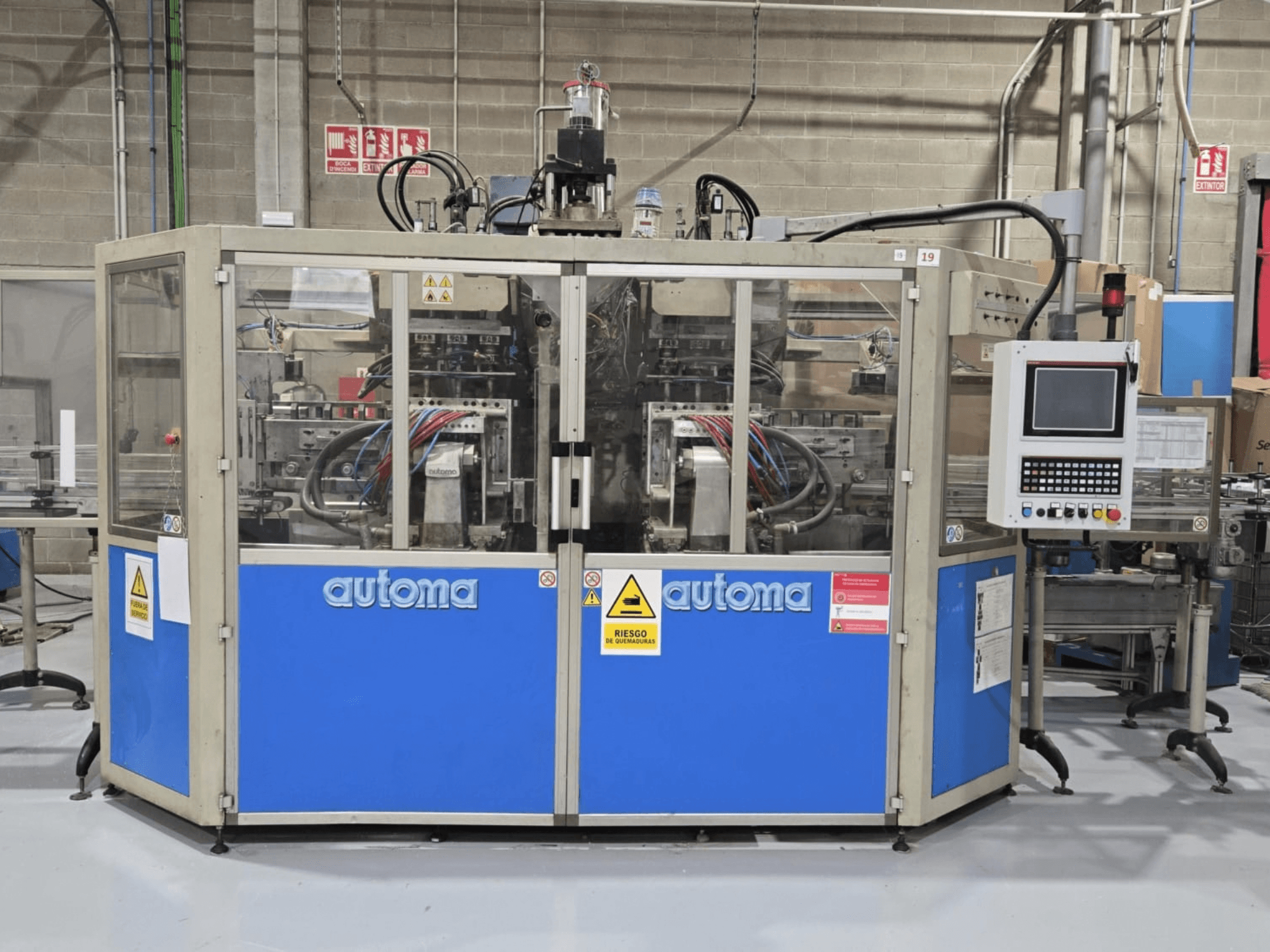 AUTOMA APEX AT500D şişirme kalıplama makinesinin, kontrol paneli ve çeşitli hortumların görüldüğü ön görünümü; makine bir fabrika ortamında yer almaktadır.