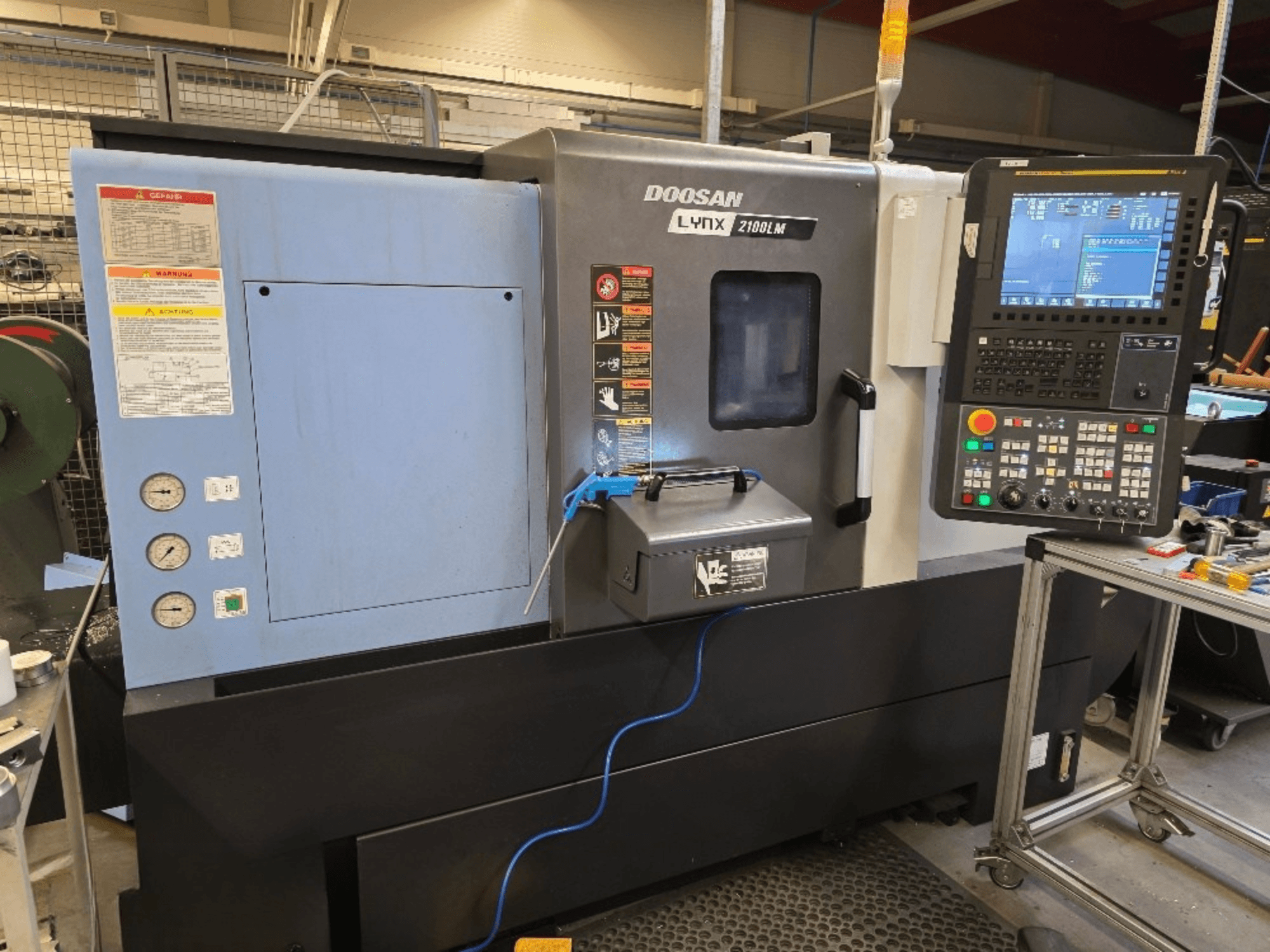 Doosan Lynx 2100LM CNC torna tezgahı, önden görünüm, kontrol paneli, basınç göstergeleri ve güvenlik uyarıları.