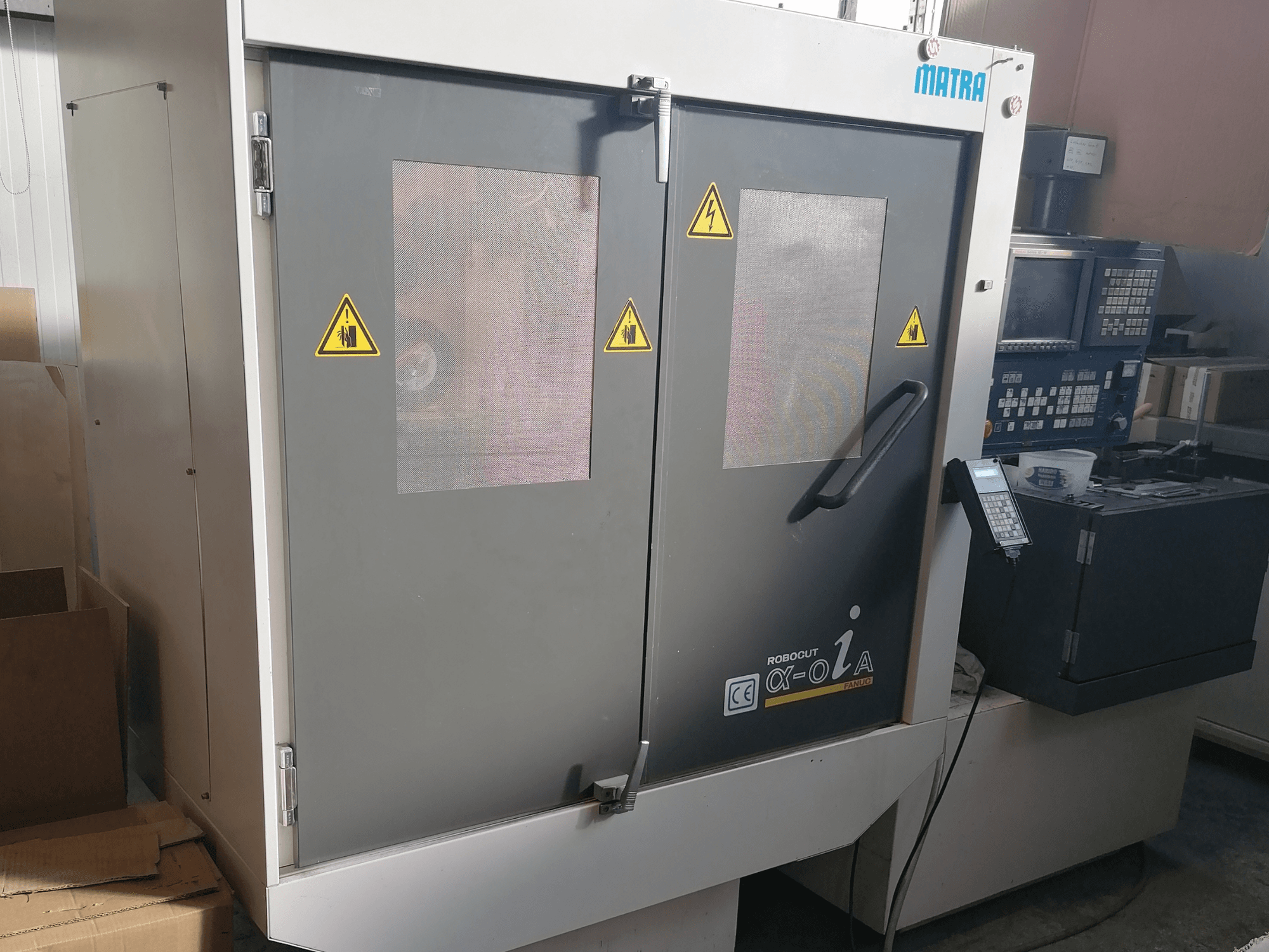 Matra'nın Robocut α-oZα makinesi, güvenlik uyarılarını, kontrol panelini ve kafes pencereli çift kapıyı gösteren önden görünüm.