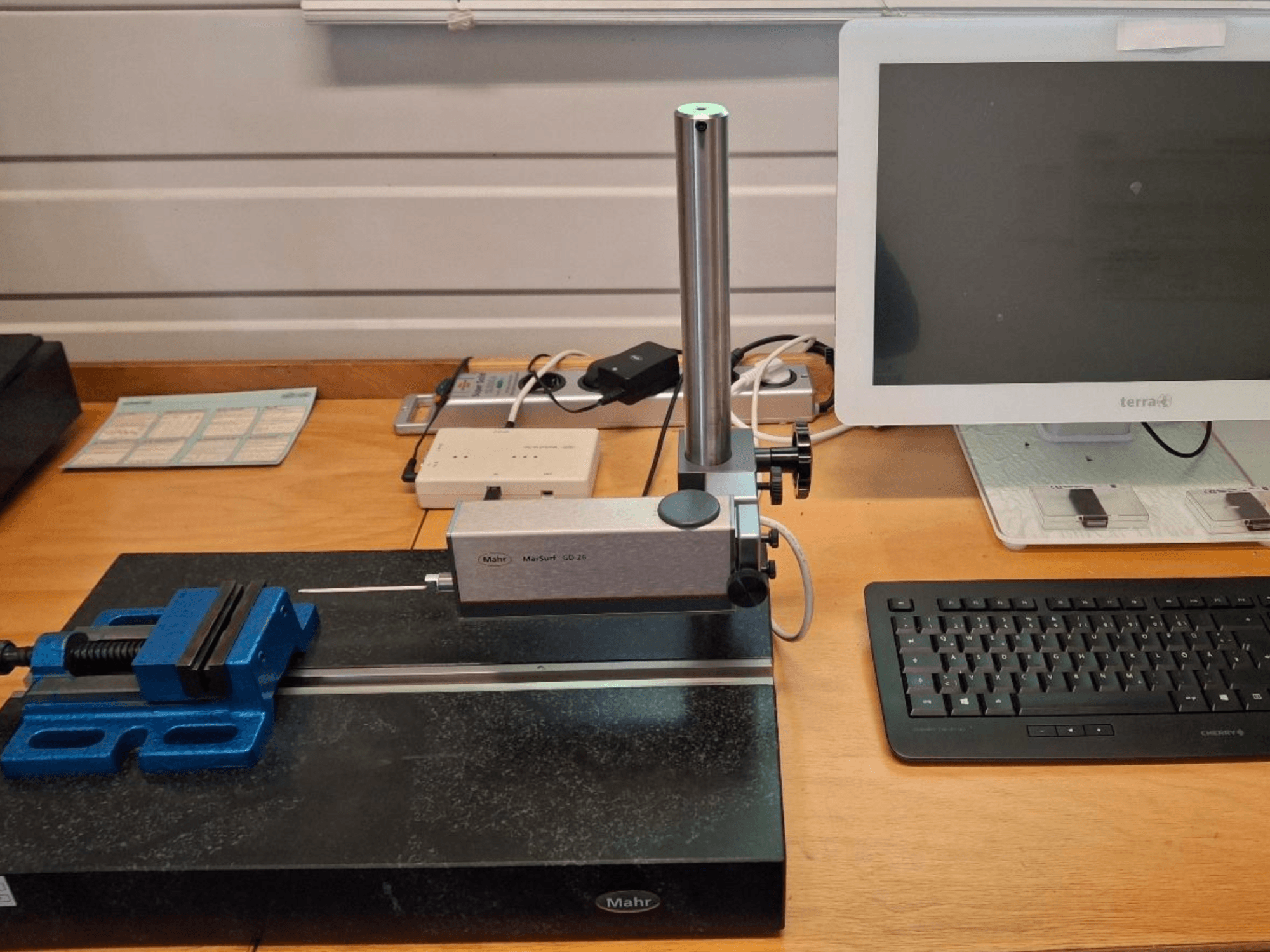 MAHR MARSURF GD 26 CMM, bir ölçüm probu, bir mavi mengene ve bir bilgisayar monitörü içeren bir çalışma tezgahı üzerinde. Önden görünüm.