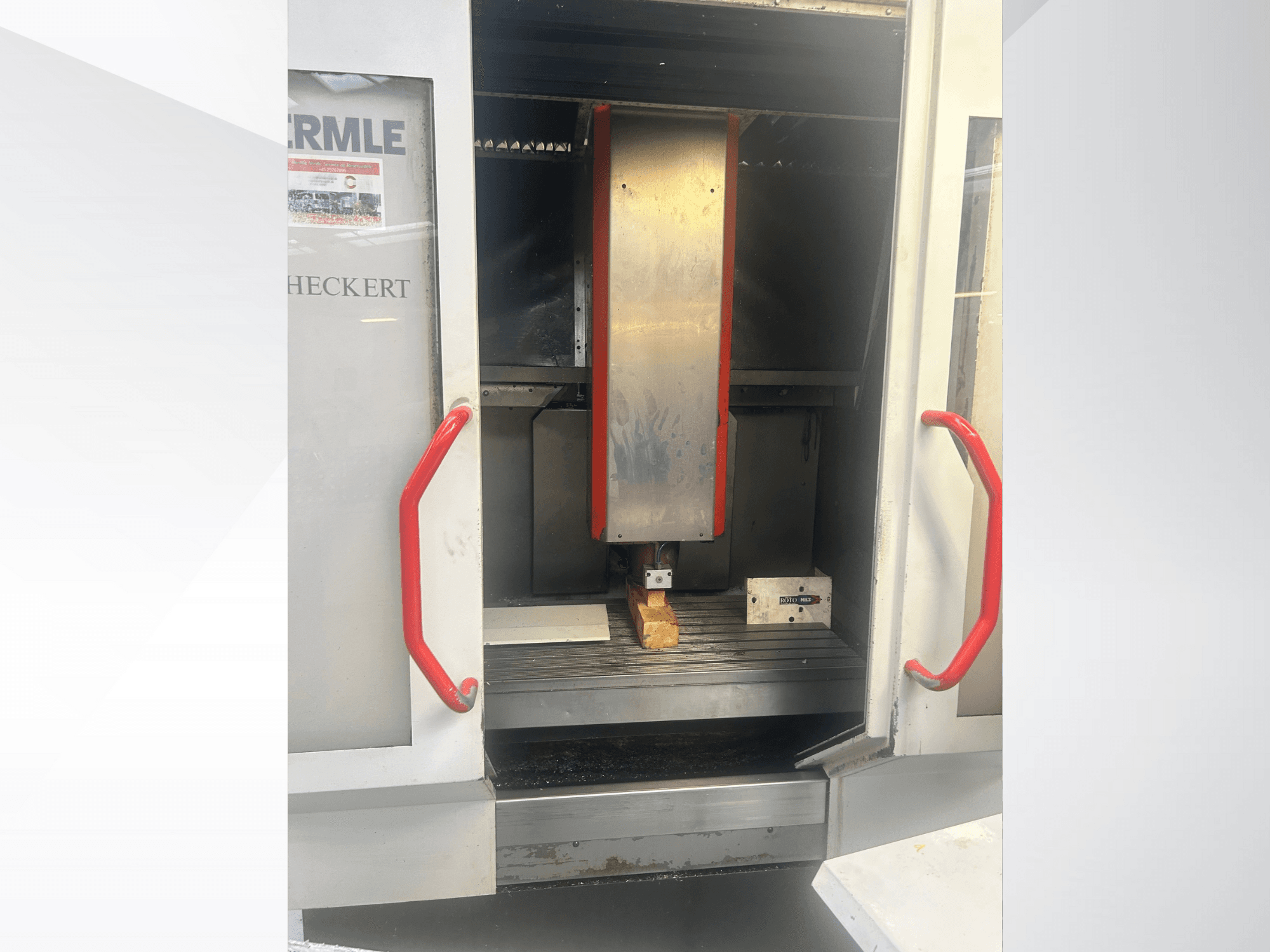 Hermle CNC makinesinin önden görünümü, metalik iç kısım, kırmızı tutamaklar ve tabla üzerinde bir iş parçası.