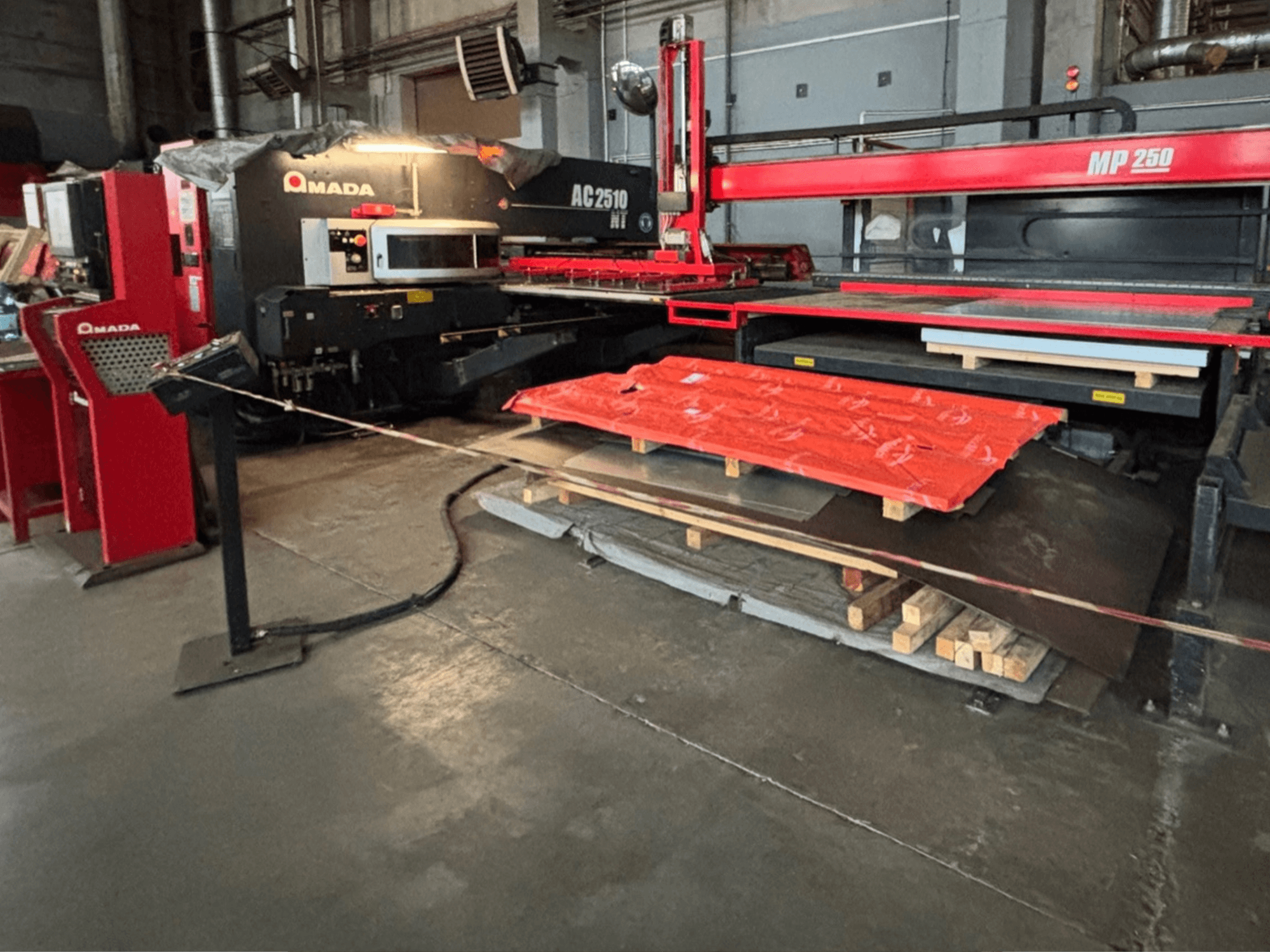 Fabrika ortamında Amada AC2510 NT lazer kesici, önden görünüm, zeminde metal levhalar ve paletler bulunan ağır makineler.
