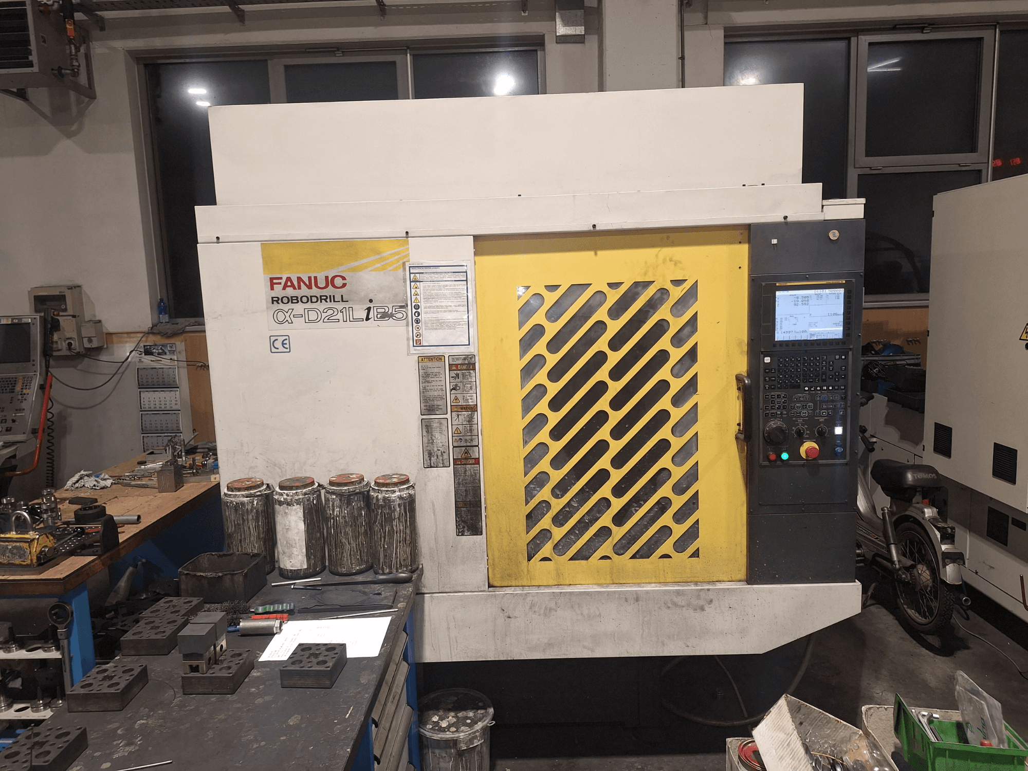 FANUC Robodrill α-D21LiB5'in önden görünümü, kontrol panelini, sarı ızgarayı ve bir çalışma tezgahı üzerindeki yakındaki aletleri gösterir.