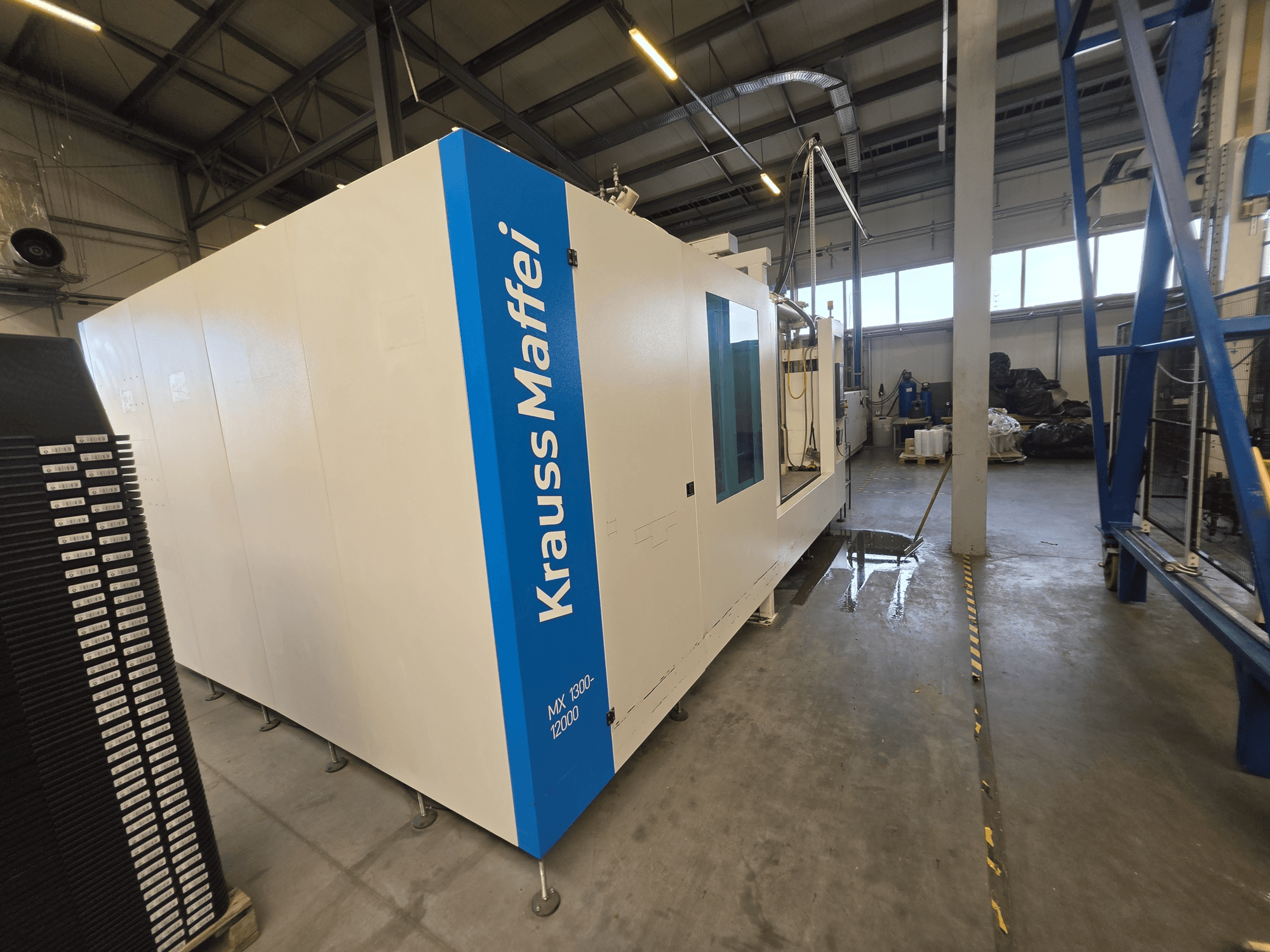 Krauss Maffei MX 1300-12000 hidrolik enjeksiyon kalıplama makinesinin yandan görünümü; makinenin geniş beyaz şasisi ve mavi marka logosu göze çarpıyor.