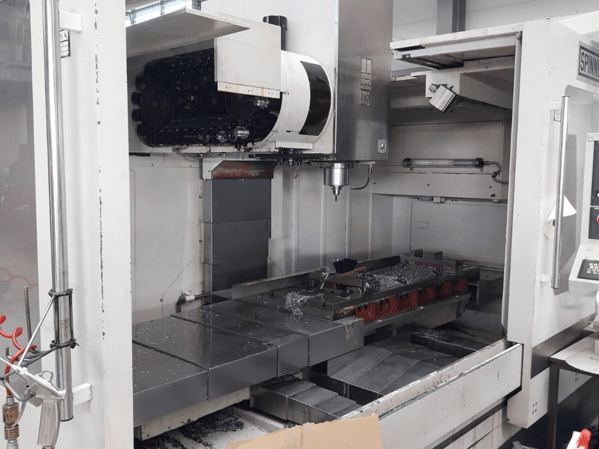 SPINNER MVC 1600 dikey işleme merkezinin açık çalışma alanını, iş milini ve tabla üzerindeki metal talaşları gösteren önden görünümü.